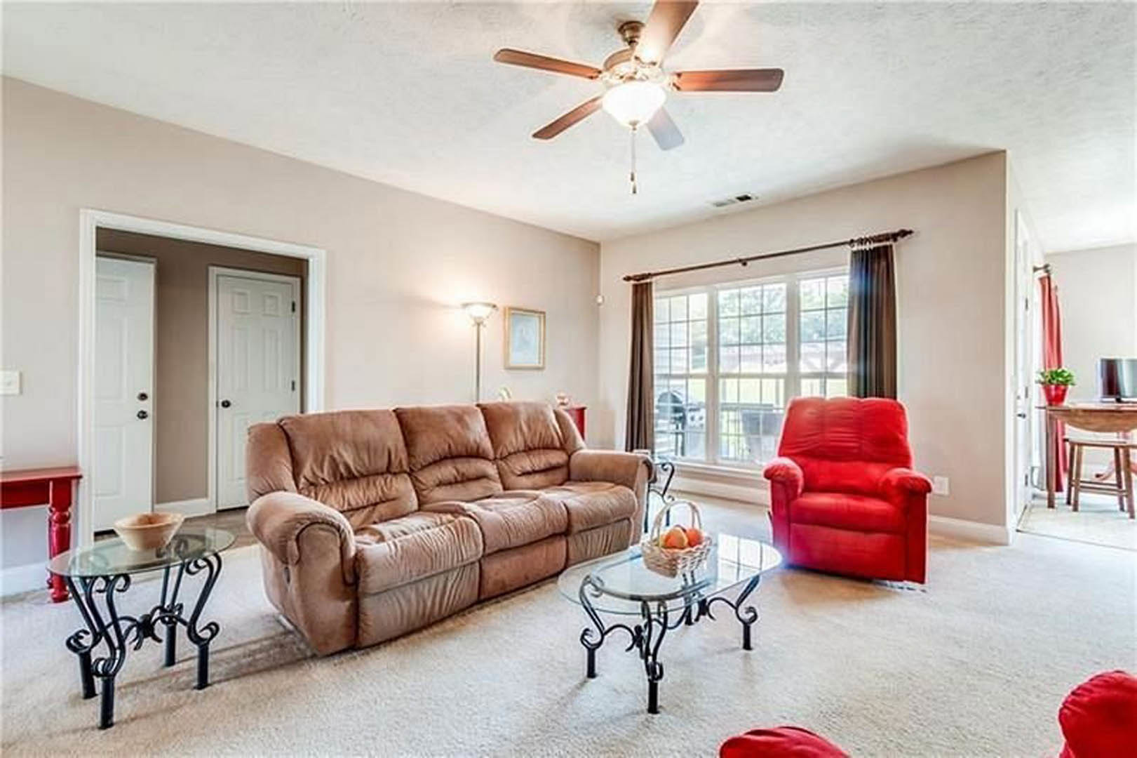 Living room with light-colored couch, red recliner chair, glass coffee table holding a bowl of fruit, neutral walls, wood flooring, ceiling fan overhead