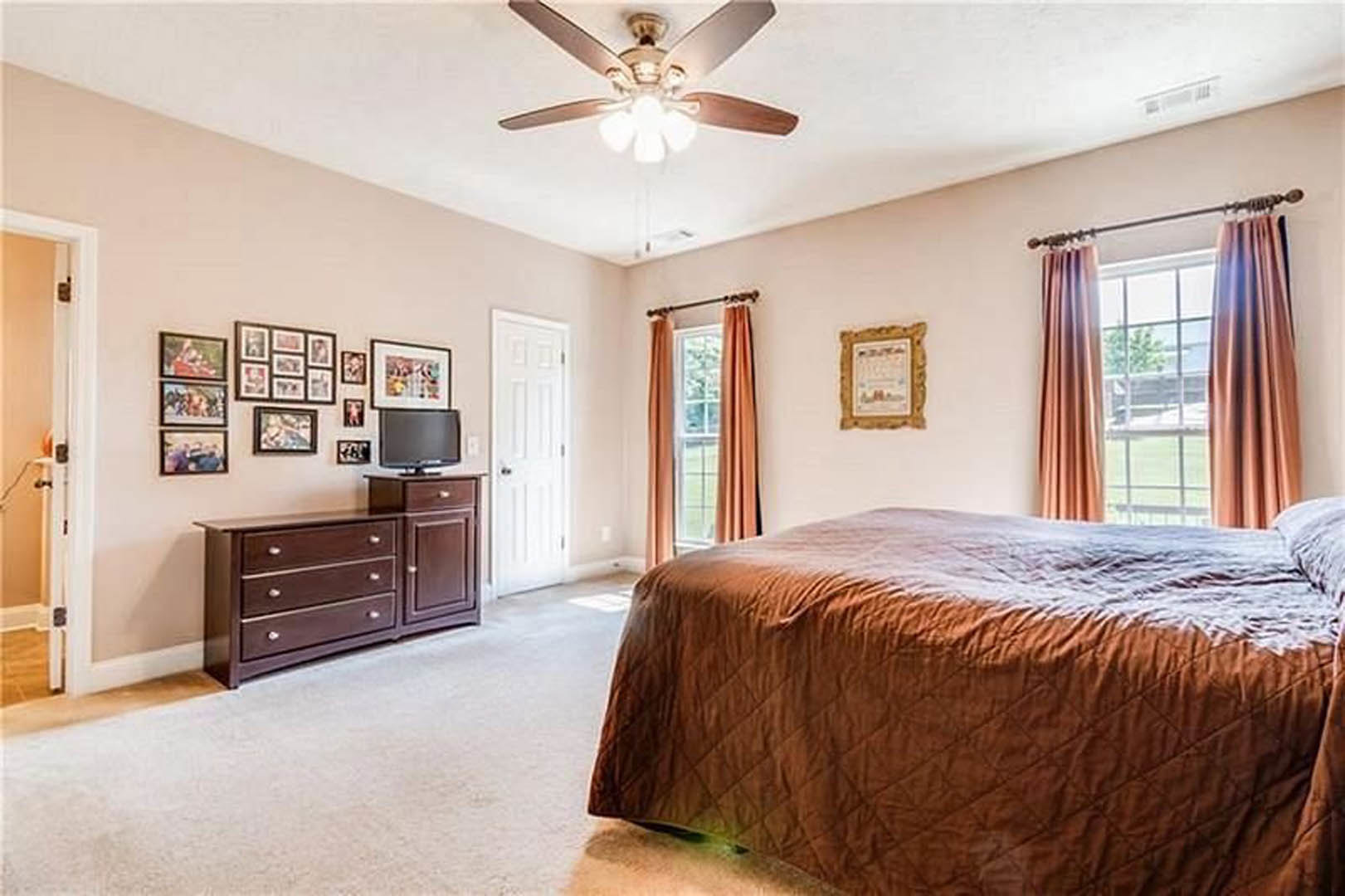 Bedroom with brown blanket on bed, ceiling fan with light fixture, window with curtains, white door with silver handle, television on wall, neutral walls and flooring
