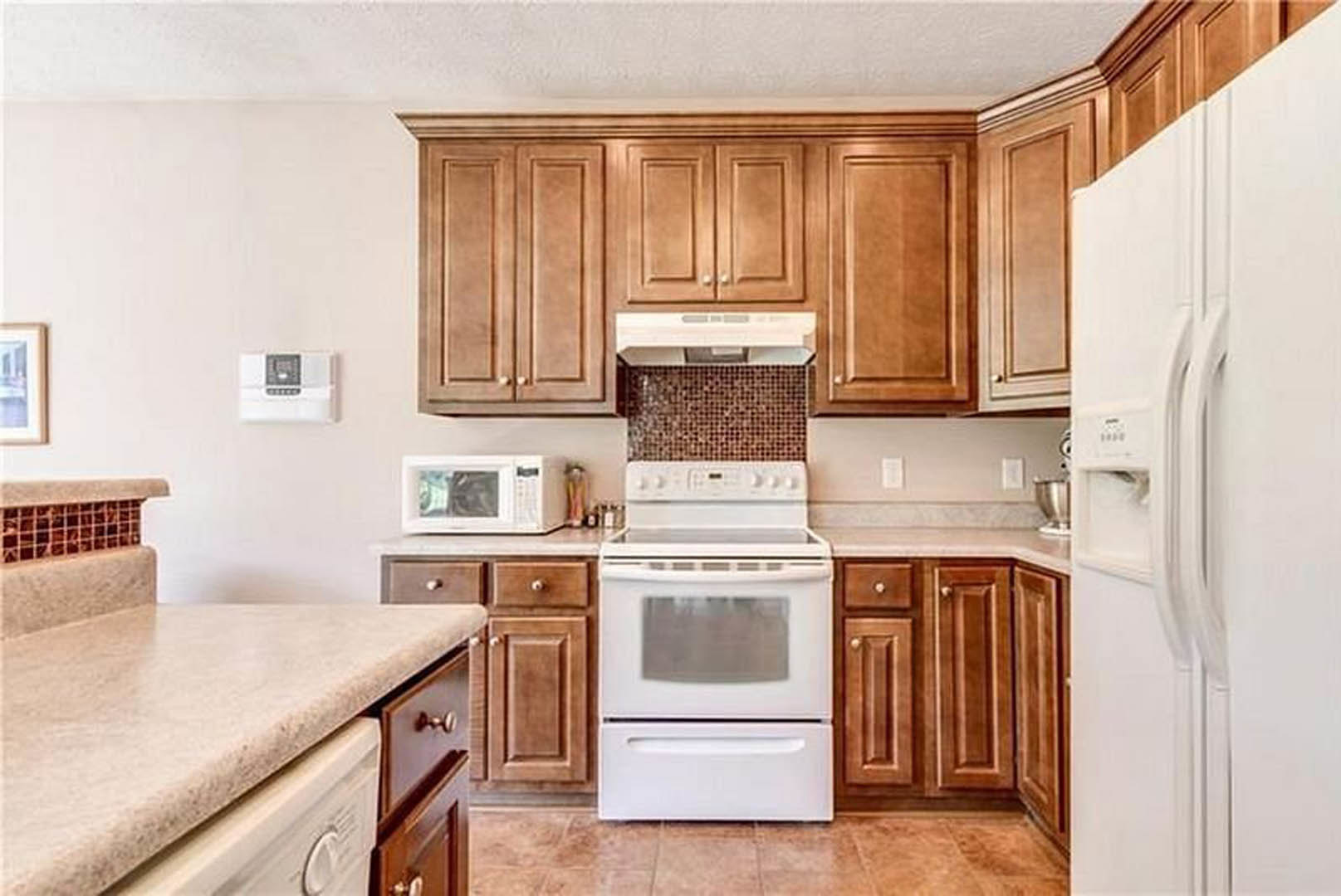 Brown wood cabinets and white appliances including refrigerator, oven, microwave, and washing machine, with a light-colored countertop in a residential kitchen.