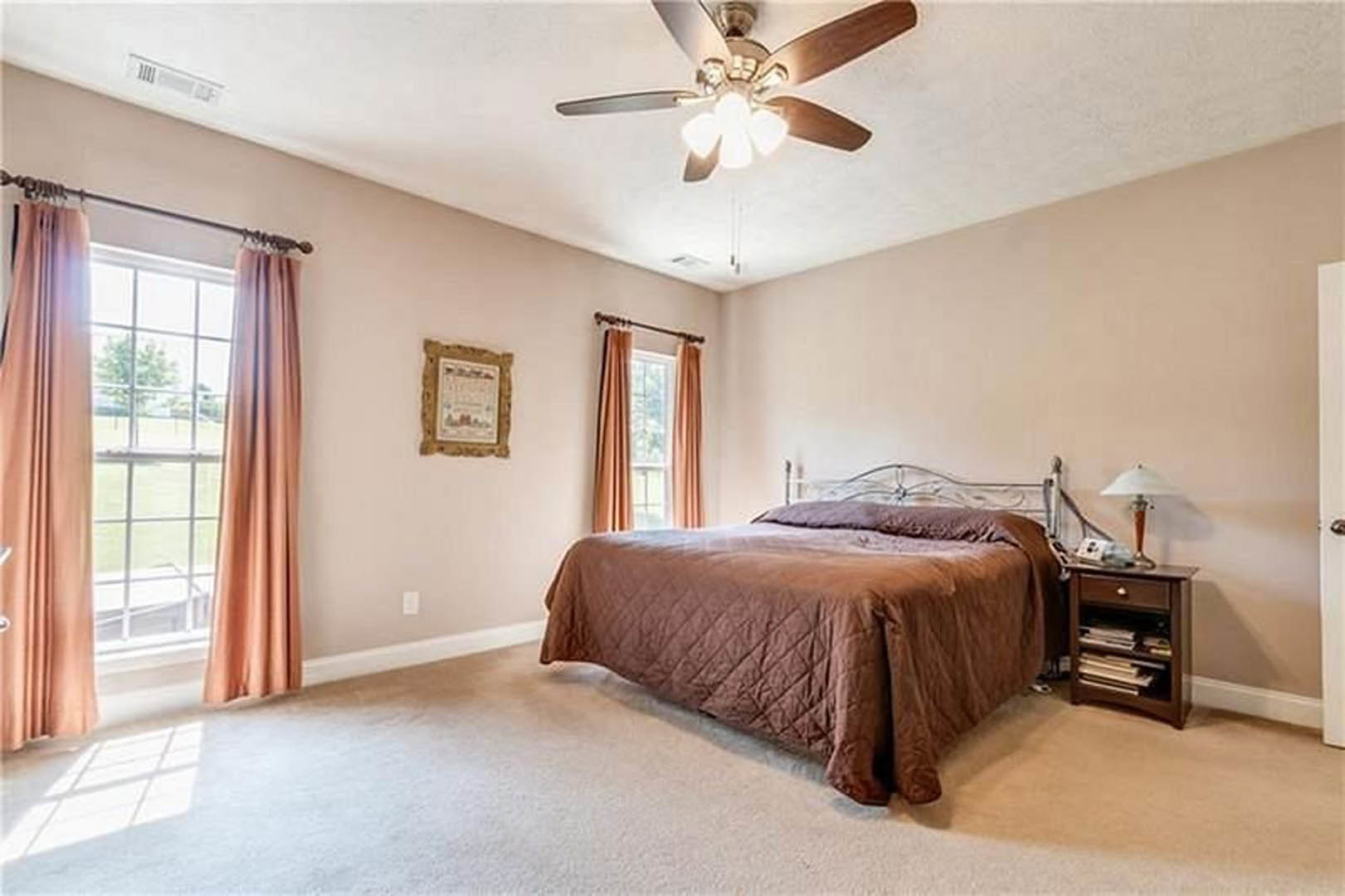 Bedroom with a bed featuring a brown blanket and pillows, ceiling fan with light fixture, window with curtains overlooking grass, wall-mounted shelf holding books, framed picture