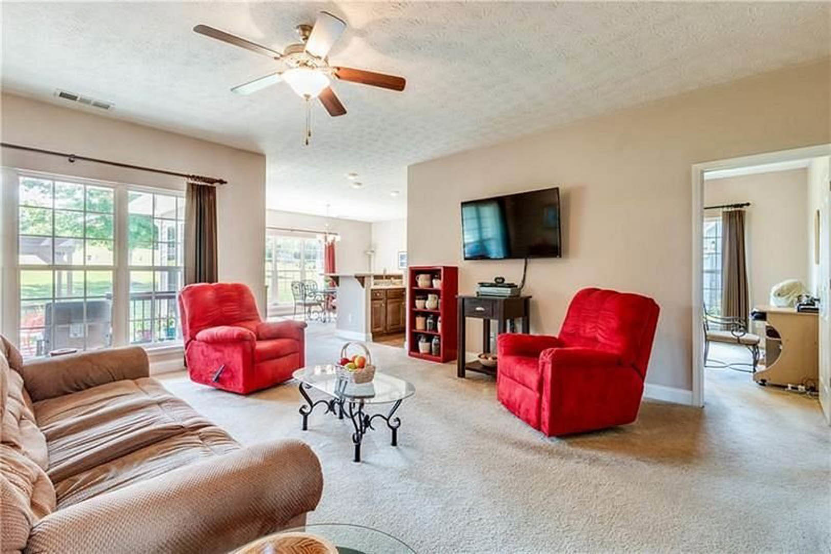 Living room with red upholstered chairs, gray couch, glass coffee table holding a fruit basket, wall-mounted television, ceiling fan with light fixture, hardwood floors, and