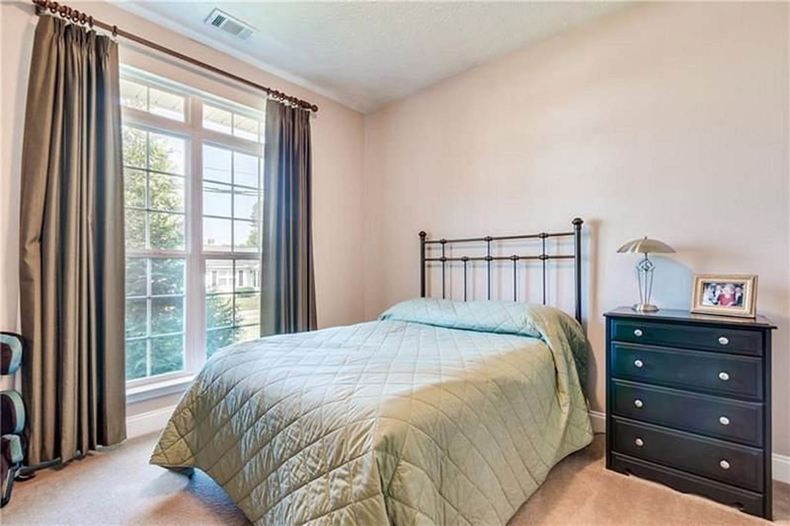 White blanket on metal headboard bed, black dresser with silver knobs, metal-shaded lamp, window with curtains, blurry family photo on wall