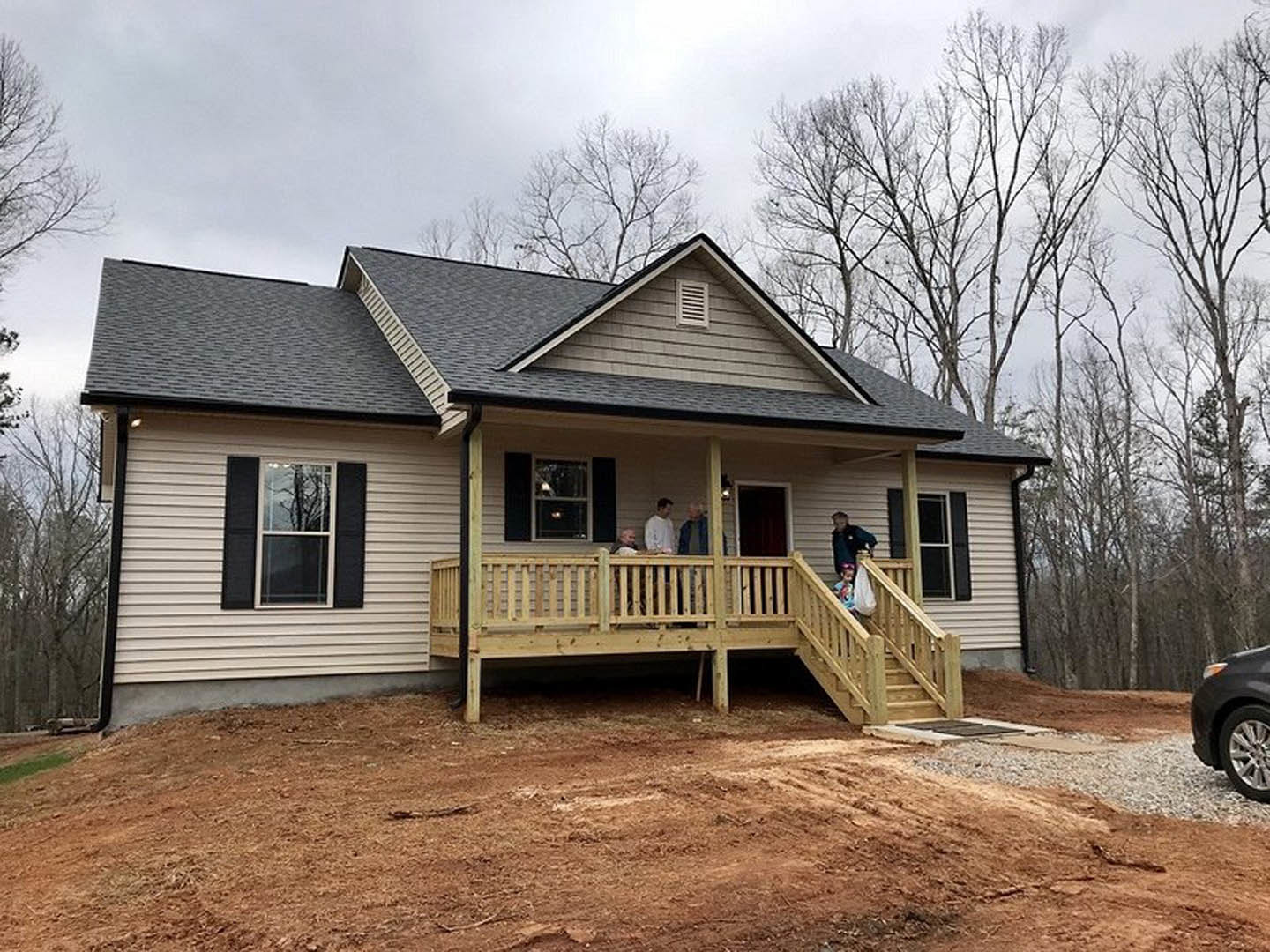 White cottage-style home with wood porch, railing, and two people standing outside; large windows reflect nearby trees, light gray siding, and cloudy sky overhead.