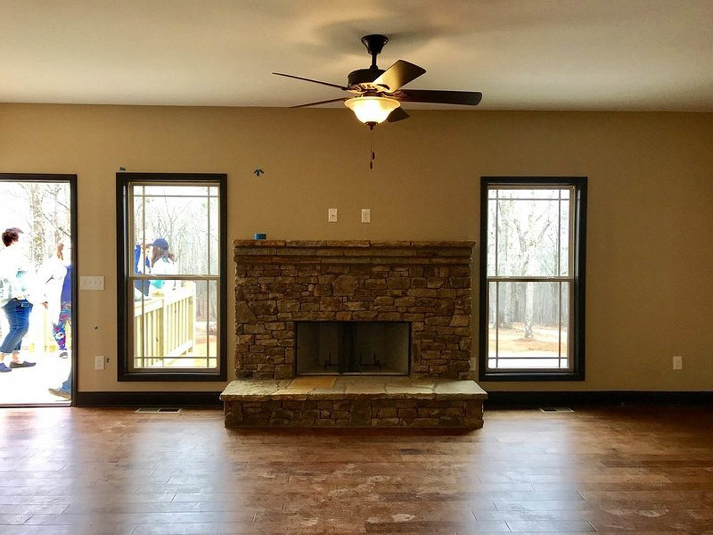 Living room with stone fireplace, ceiling fan with light, hardwood flooring, large window overlooking forest, two people near window, neutral wall finishes