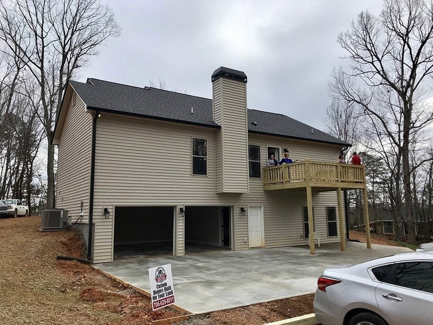Two-story home with attached garage, white car parked in driveway, people gathered on upper deck, large windows, wood siding, and surrounding trees.