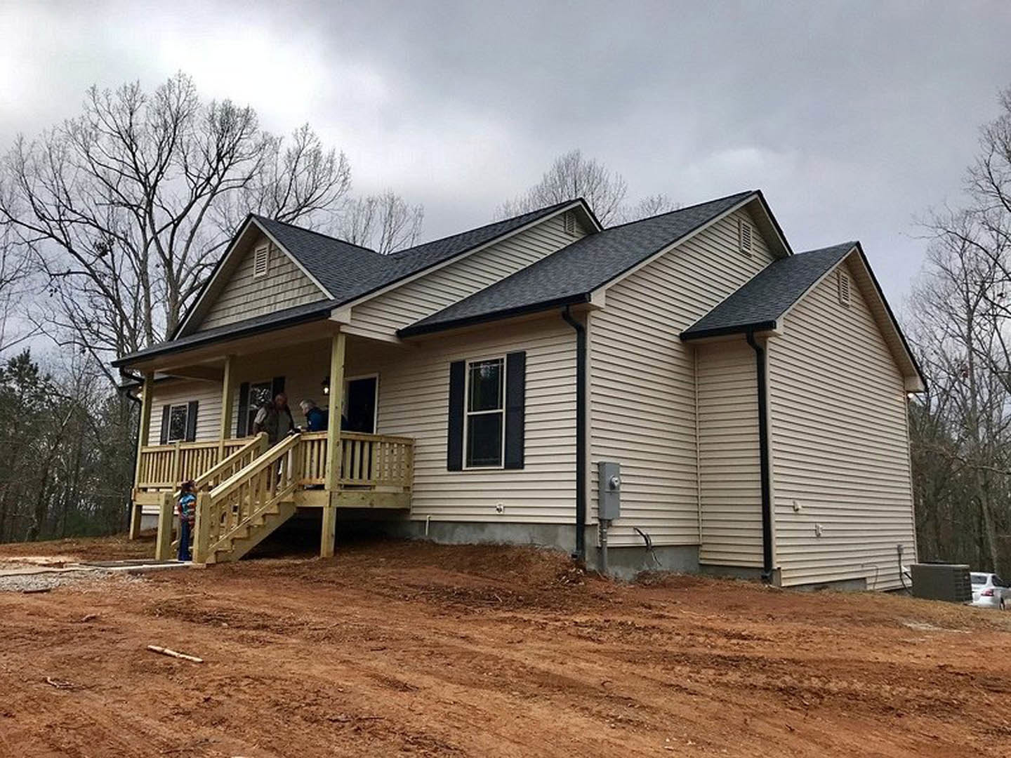 Two-story house with covered porch, wooden stairs and railings, white-framed windows, people standing near entrance, white car parked on dirt driveway, trees and cloudy sky in