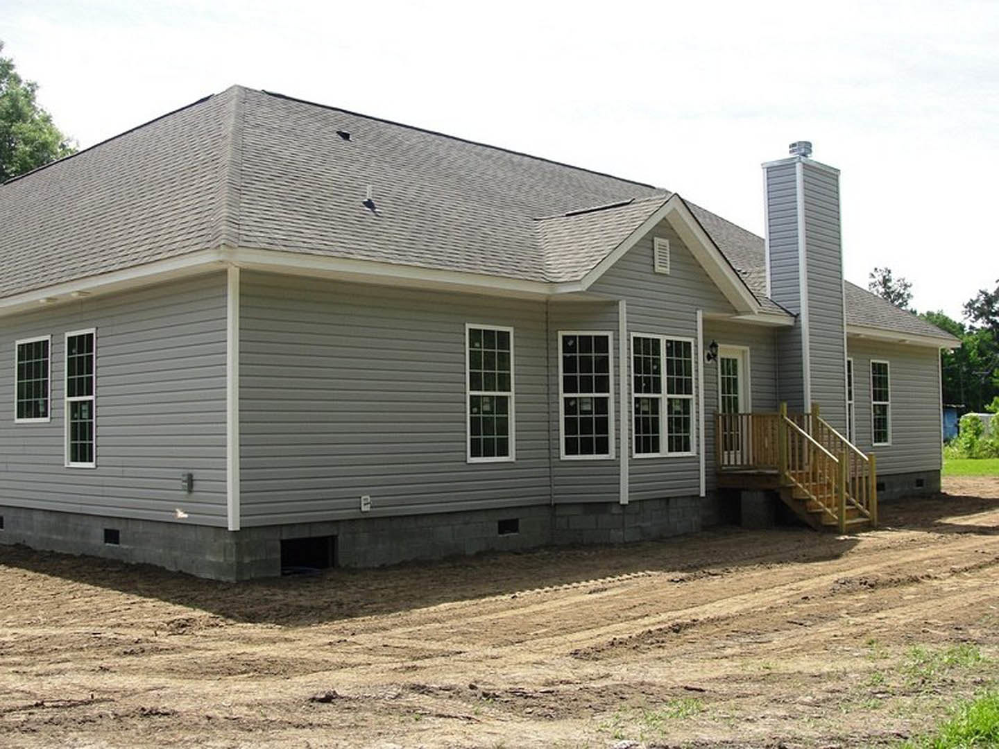 Grey cottage-style home with multi-pane windows, wood deck and stairs, surrounded by trees and a dirt road