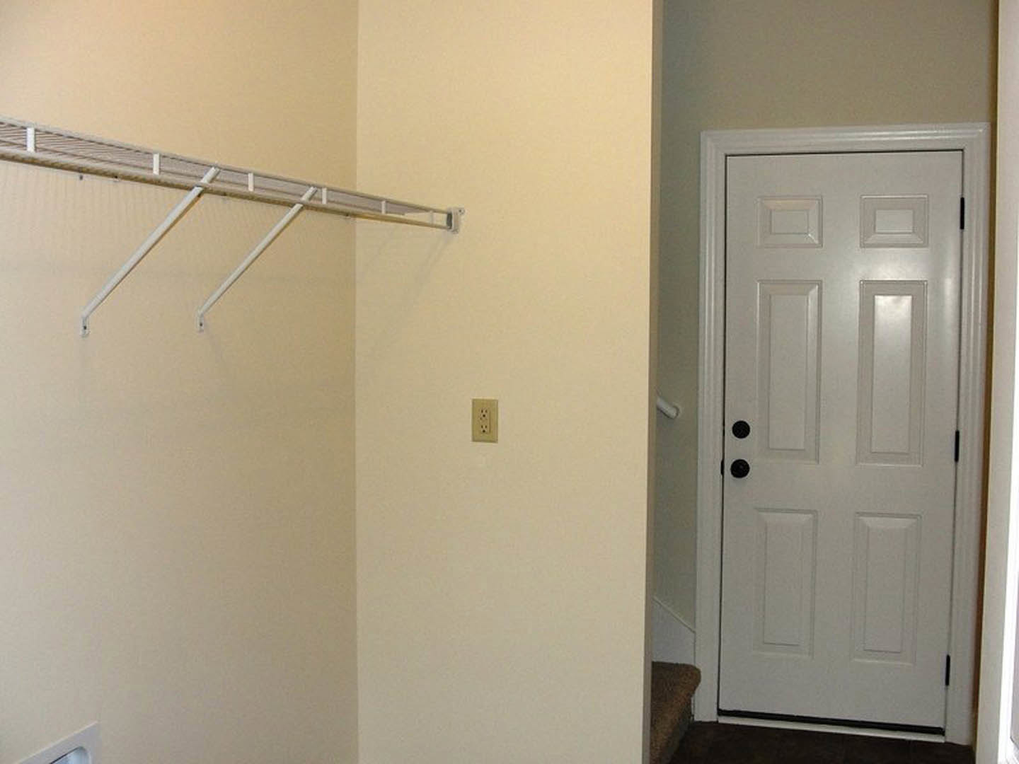 White paneled door with black knobs set in a smooth white plaster wall, electrical outlet visible nearby, white metal rod mounted above, light-colored flooring.