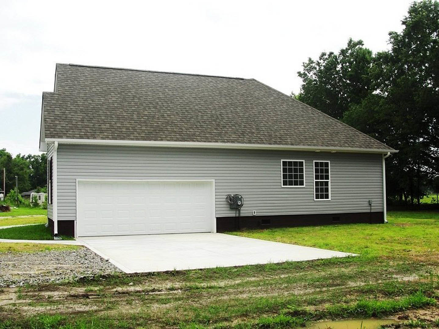 White garage door with horizontal panels, concrete patio bordered by green grass, white-framed window, light siding, mature trees in background