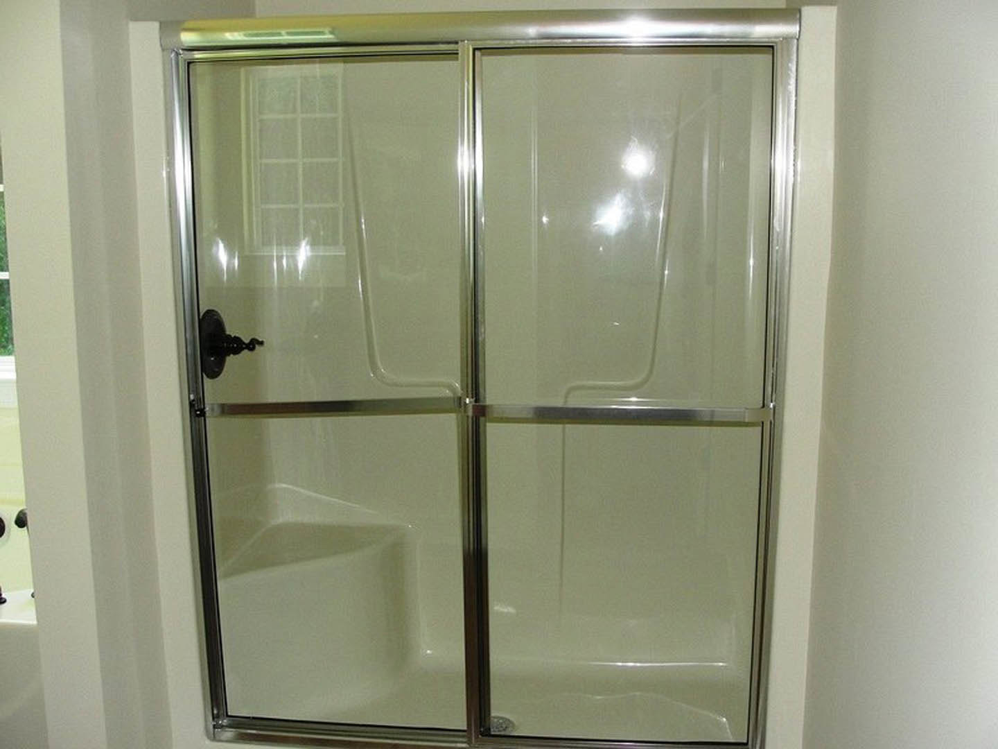 Frameless glass shower enclosure with chrome door handle, white tile walls, and natural light reflecting on surfaces