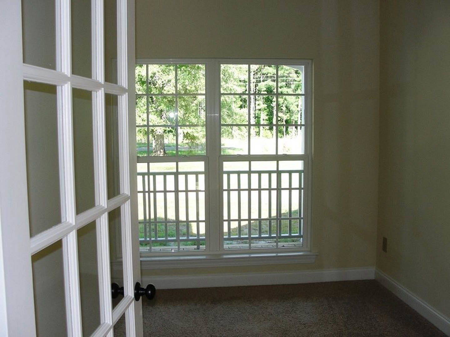 Bright room featuring a white double door opening to a balcony, large window with blinds, light-colored carpet flooring, and neutral painted walls