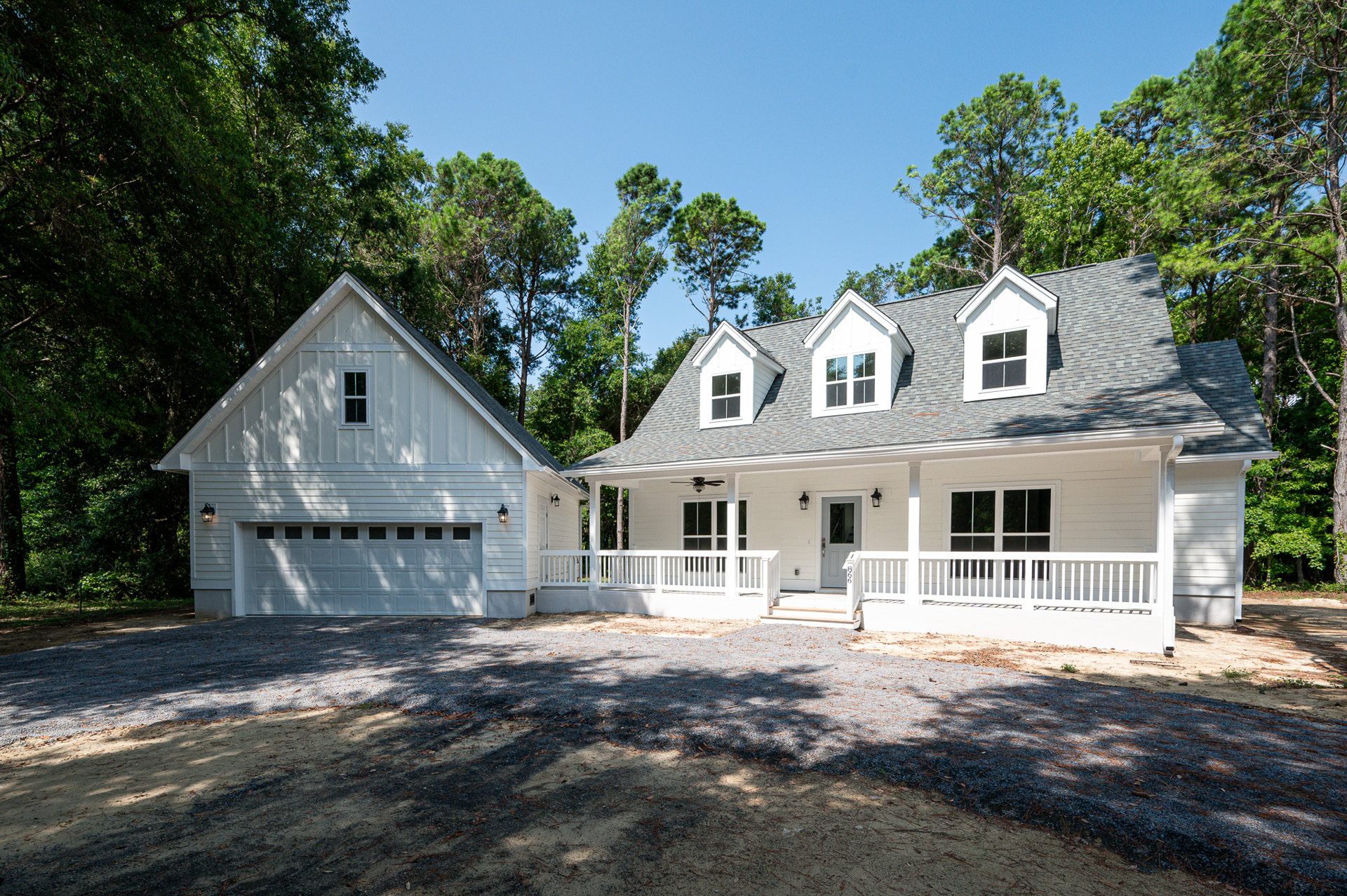 White house with attached garage, paved driveway, white fence and wall, large window reflecting trees, dirt and gravel area bordered by white railing.