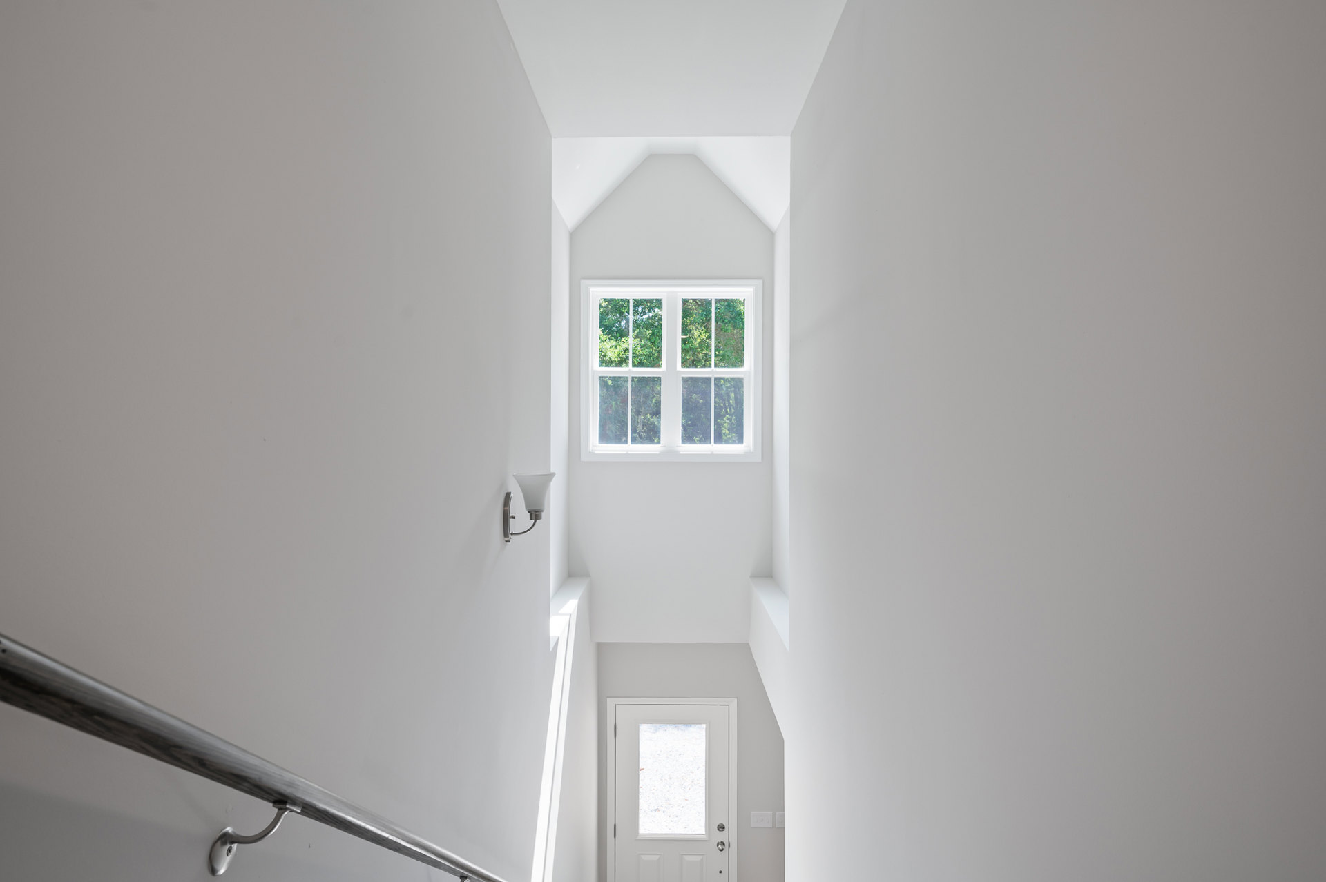 Hallway with smooth plaster walls, metal handrail, white door with glass panel, large window revealing leafy trees outside, natural daylight illuminating interior.