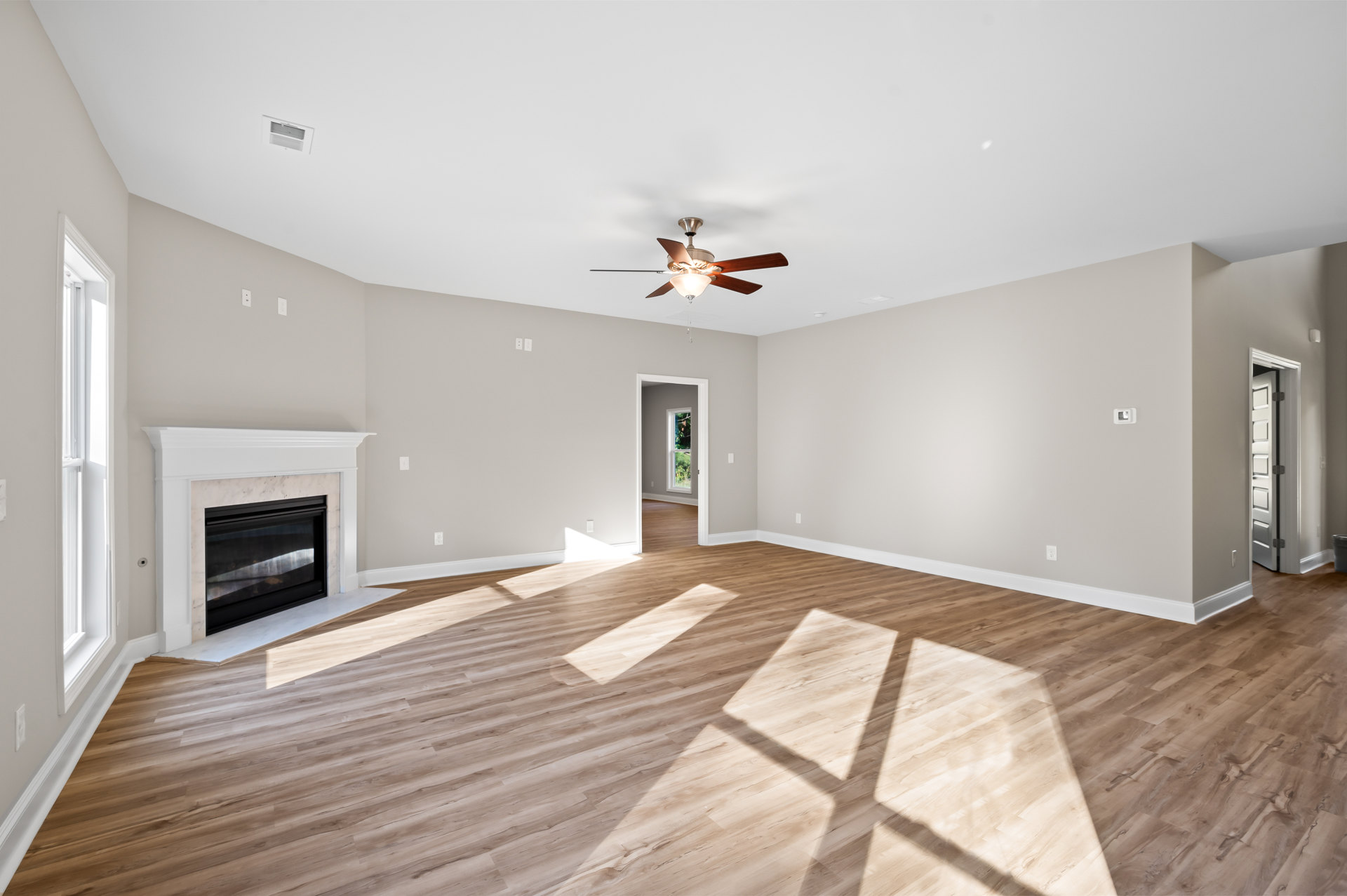 Living room with laminate wood flooring, white ceiling with vent and ceiling fan, glass-door fireplace with white mantle, open doorway leading to adjacent space
