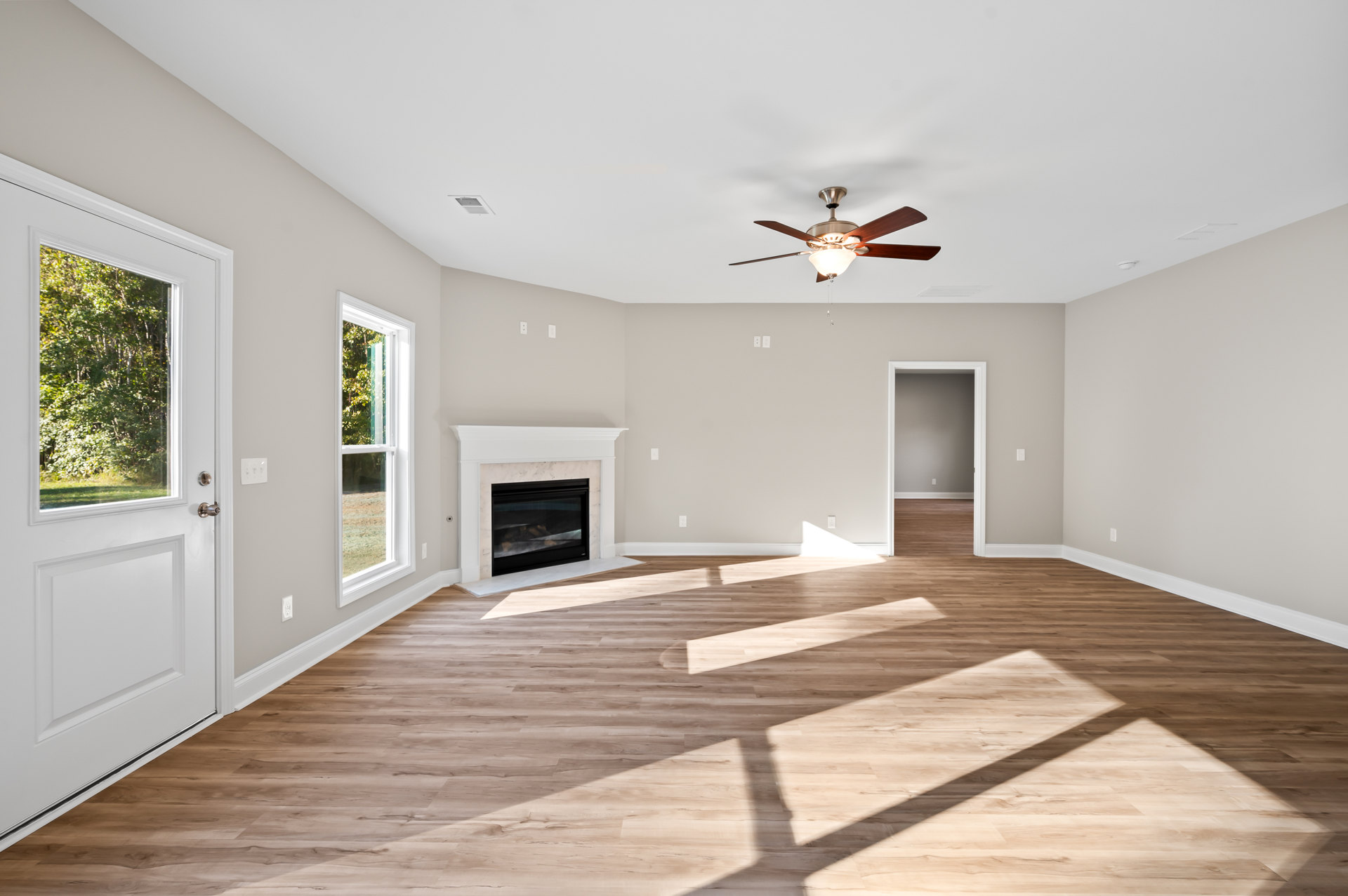 Living room with wood flooring, central fireplace with glass door and white mantel, ceiling fan with light, large window framed in white molding