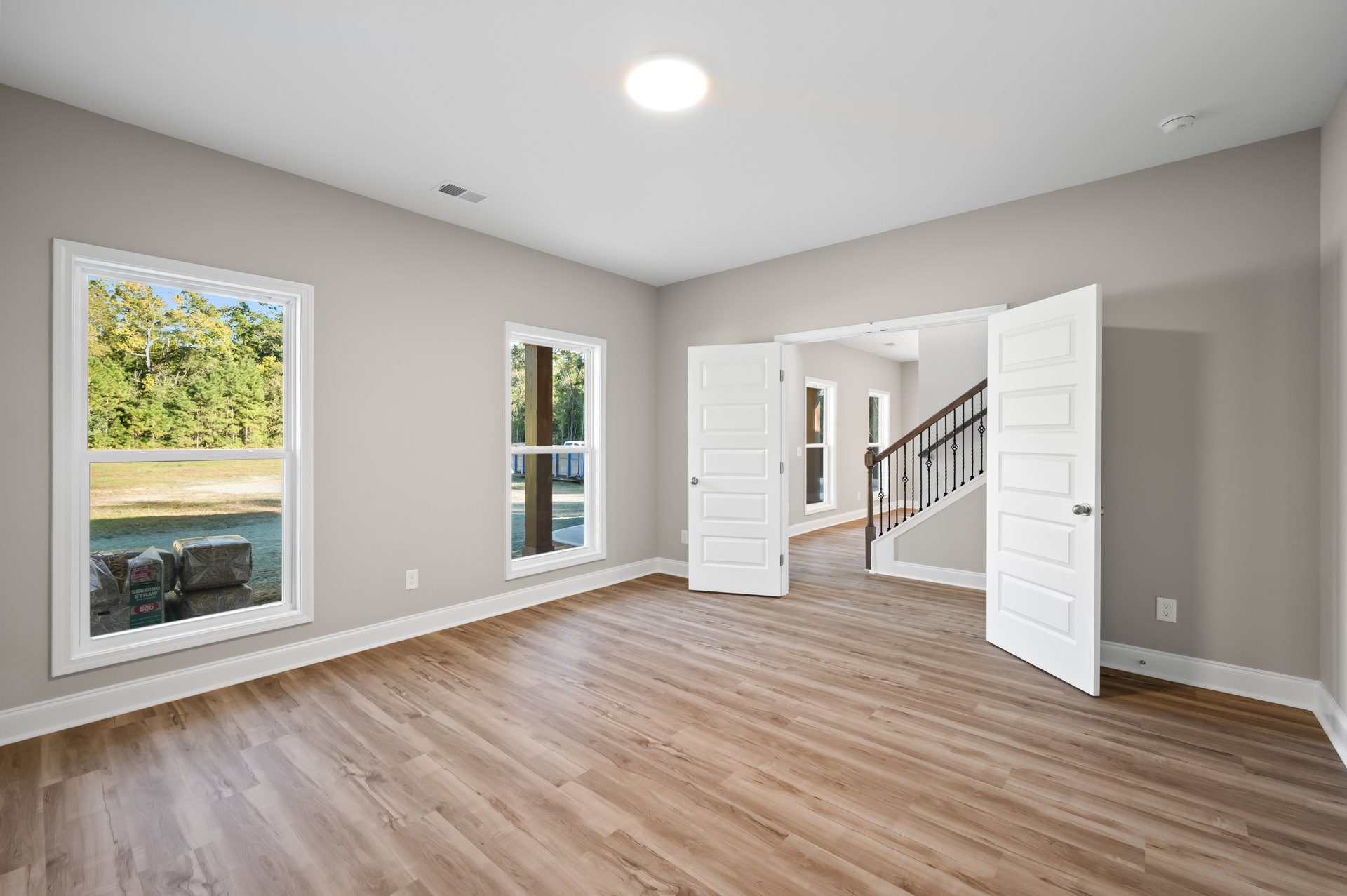 Wood flooring in a bright room featuring open white doors with silver knobs, a staircase with white risers, and windows overlooking leafy trees and stacked firewood.