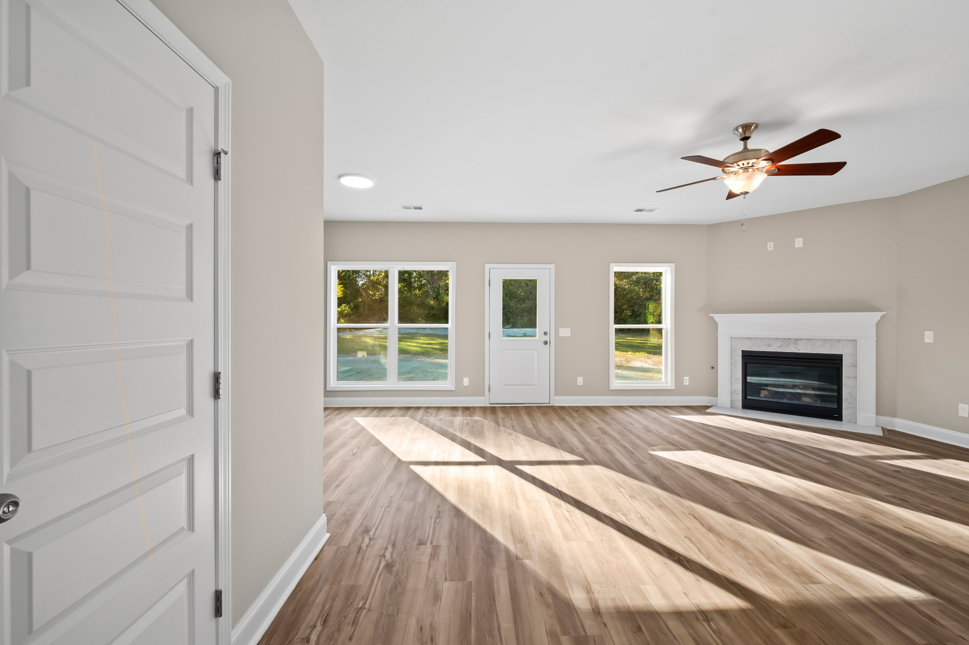 Living room with wood laminate flooring, central fireplace with glass front, ceiling fan and light fixture, white door with window, large window overlooking grassy yard and trees.