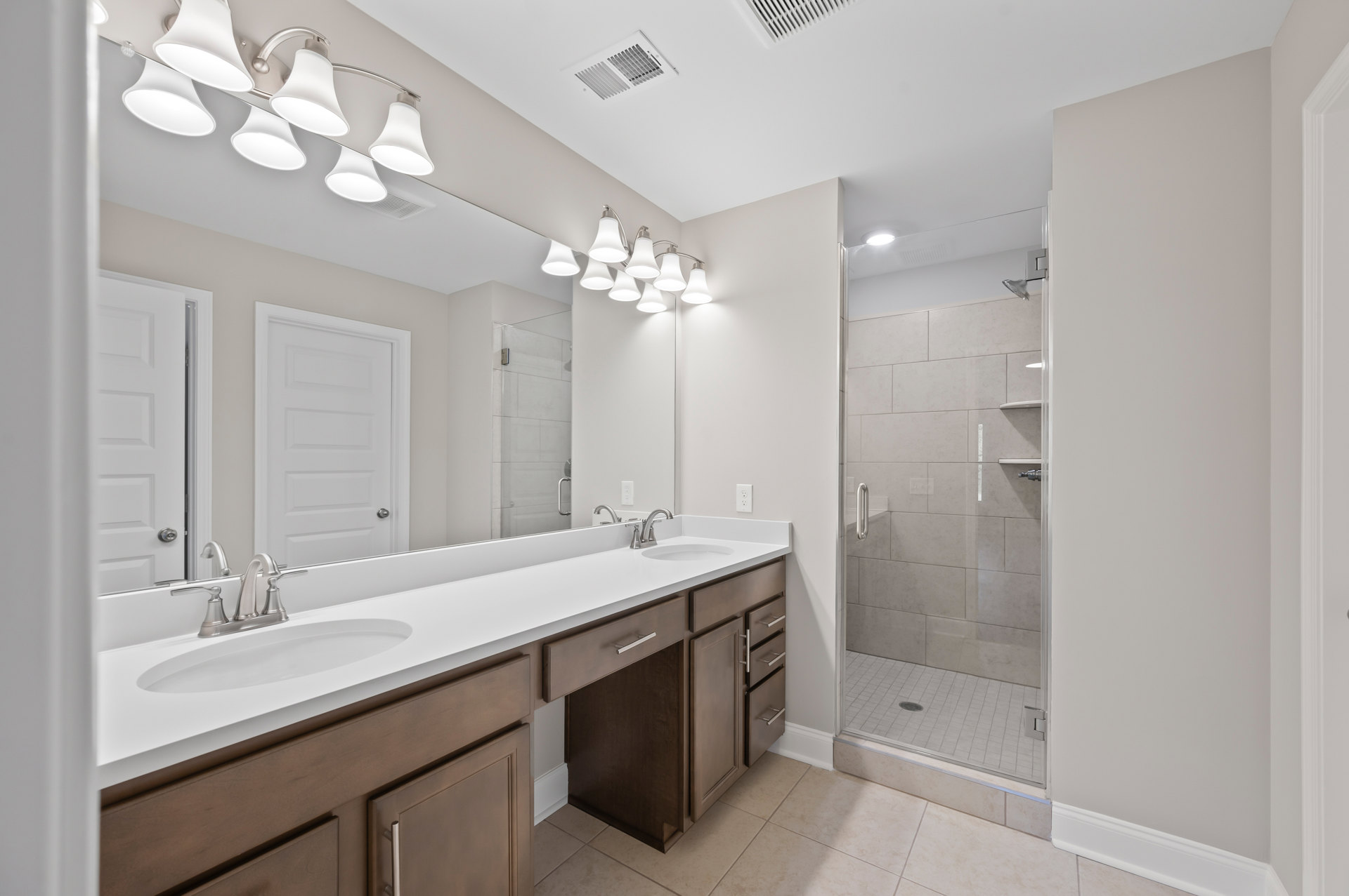 Bathroom with double vanity sinks, white tile walls, glass shower enclosure, silver hardware, and multiple lights mounted above a large mirror