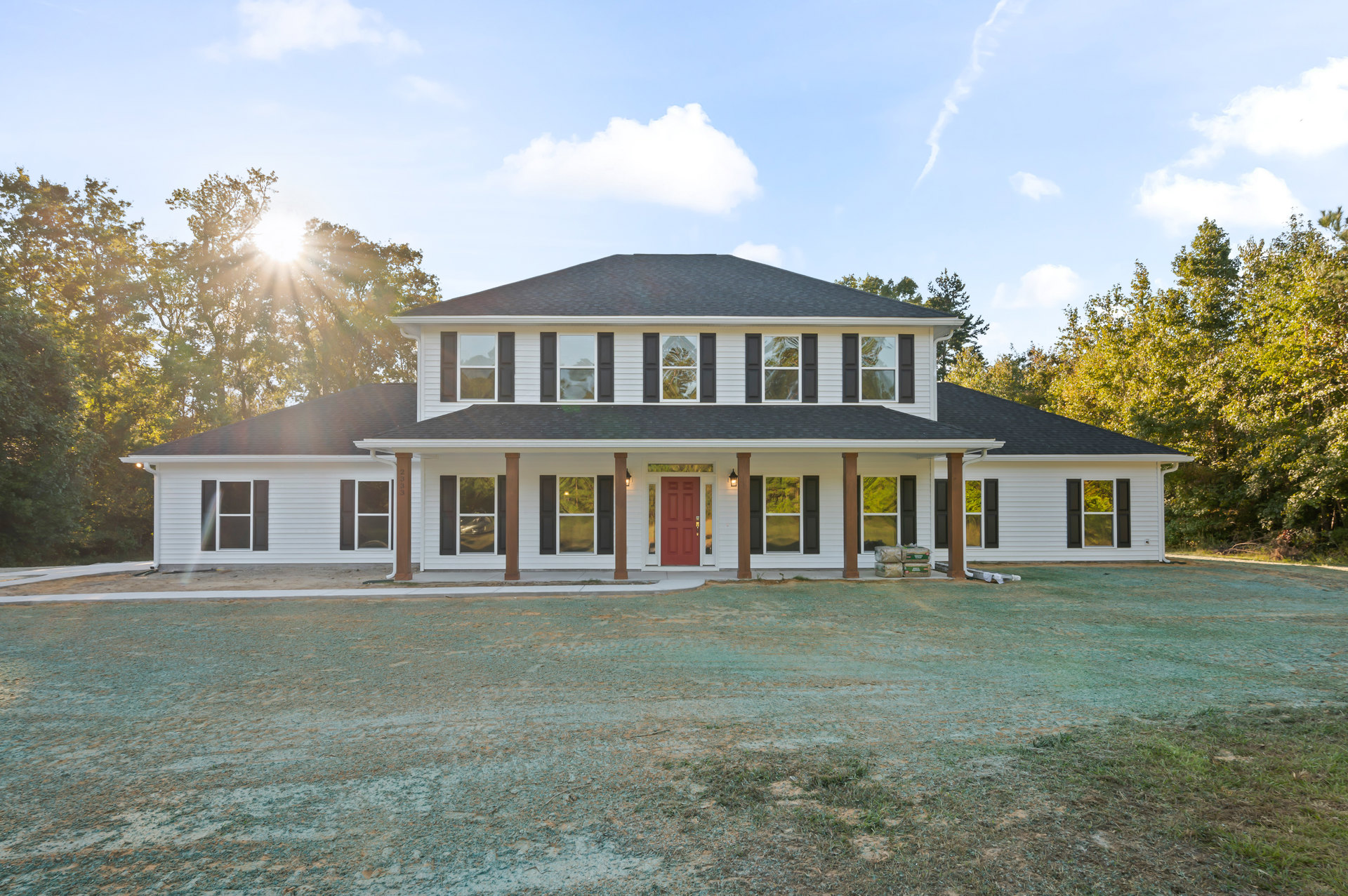 White two-story house with red front door, wide grass lawn, white-framed windows, covered porch, and blue sky with scattered clouds