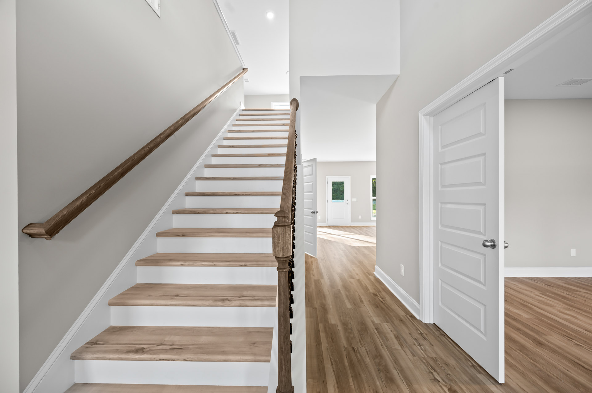 White-painted staircase with wooden steps and handrail, adjacent to a white door with glass window and silver knob, set on light hardwood flooring in a bright interior hallway.