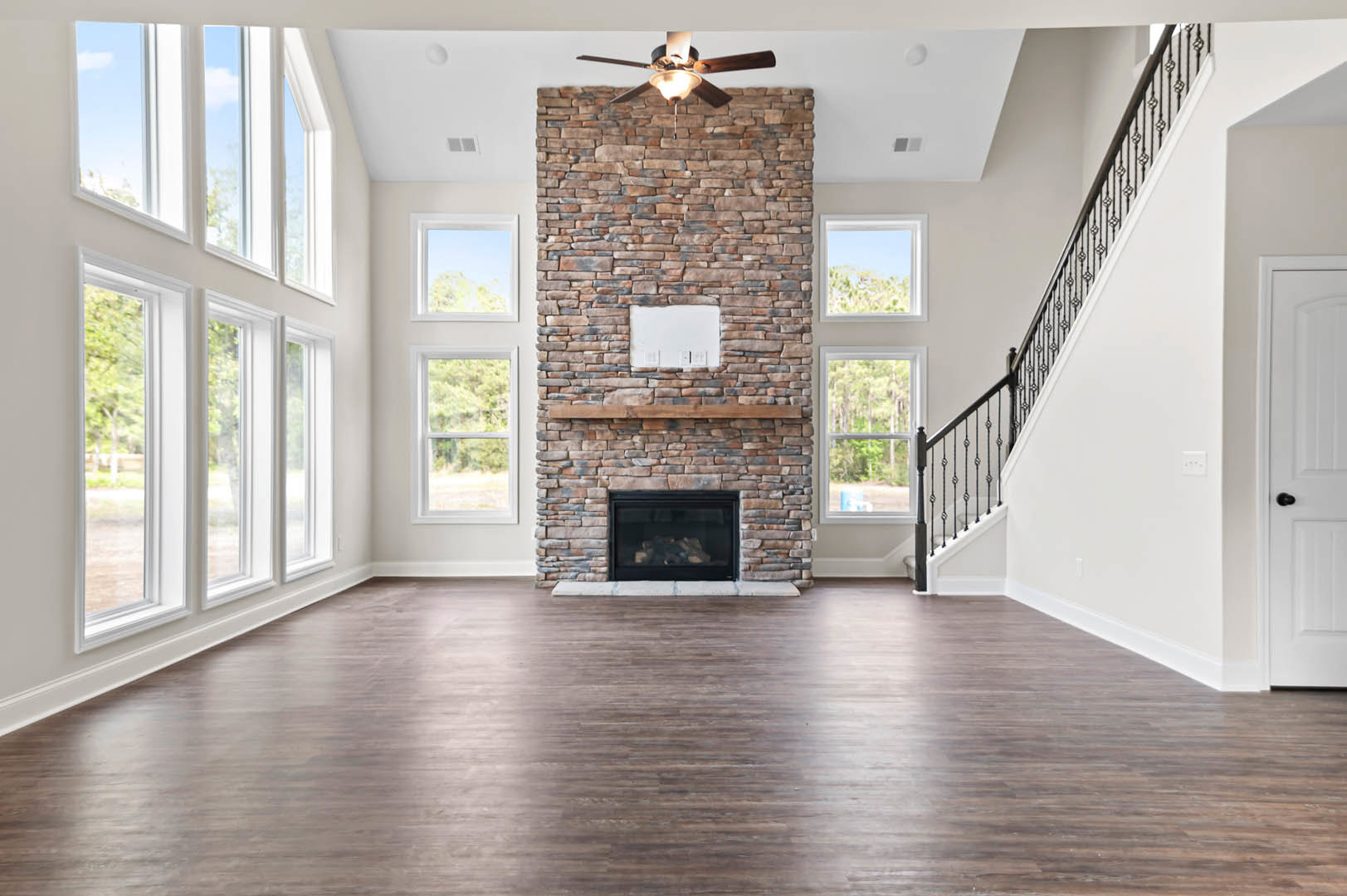 Living room featuring a stone fireplace with stacked wood, wood flooring, a staircase with wooden railing, brick accent wall with shelving, ceiling fan with light fixture, and
