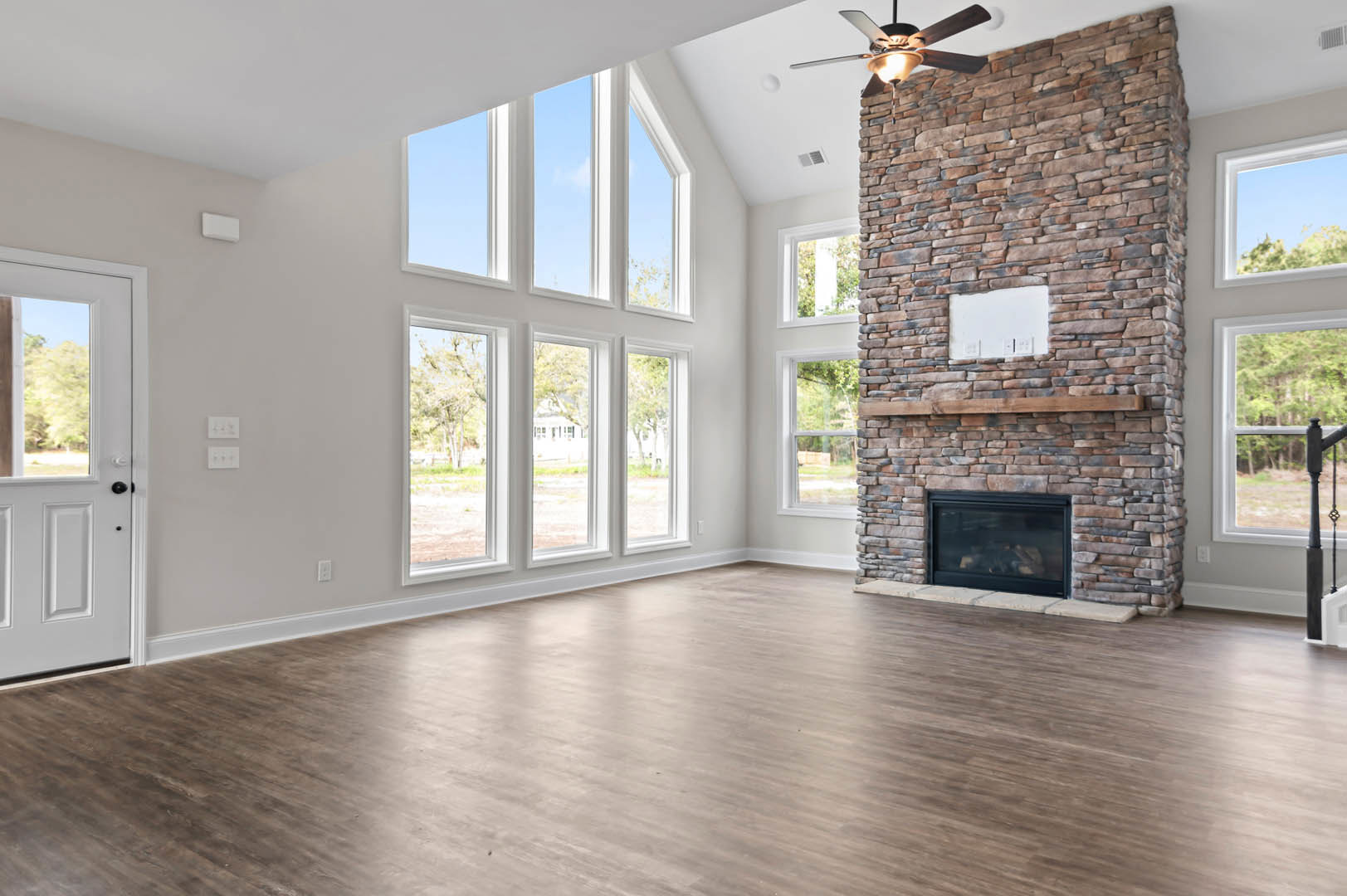 Living room featuring a brick fireplace with glass door, wood flooring, stone accent wall with built-in shelf, white walls with light switches, windows, and a ceiling fan with