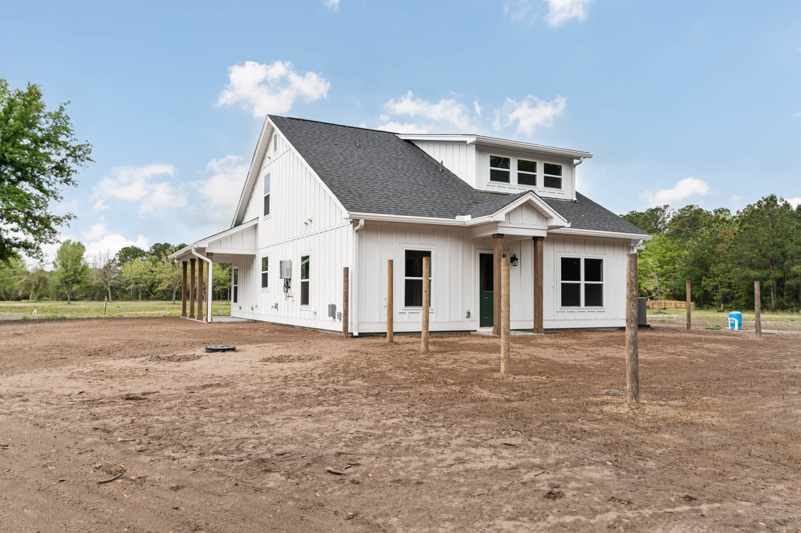 White house with wooden posts and large windows, surrounded by a dirt yard and green trees under a partly cloudy sky