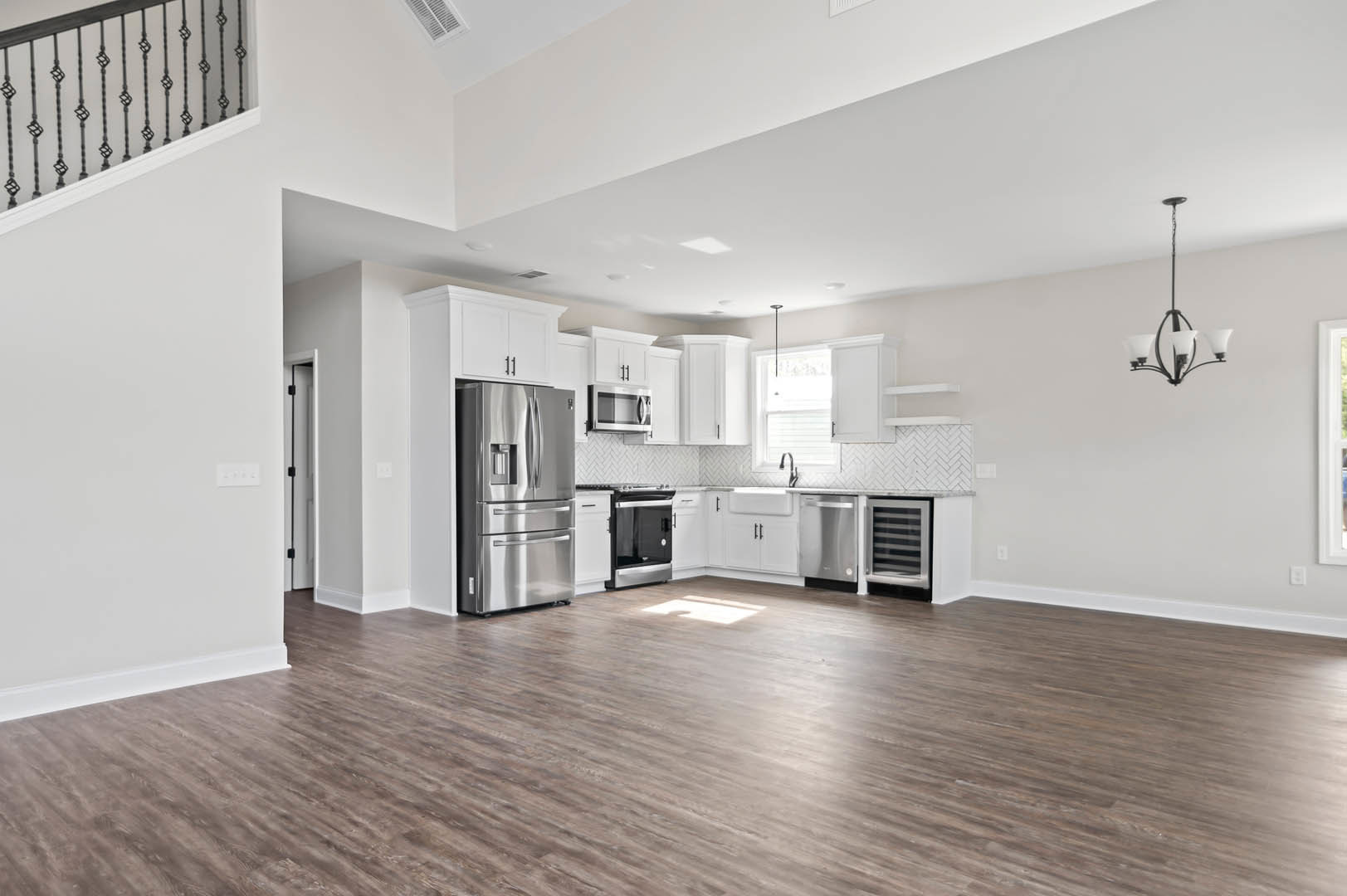 Open kitchen and living room with wood flooring, white cabinetry, stainless steel refrigerator, modern chandelier, and visible staircase.