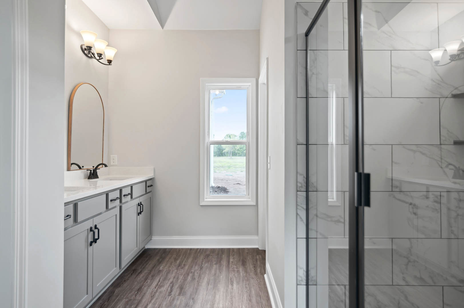 Bathroom with glass shower enclosure, white sink on wood vanity, tile flooring, window overlooking trees, wood trim, and modern light fixture