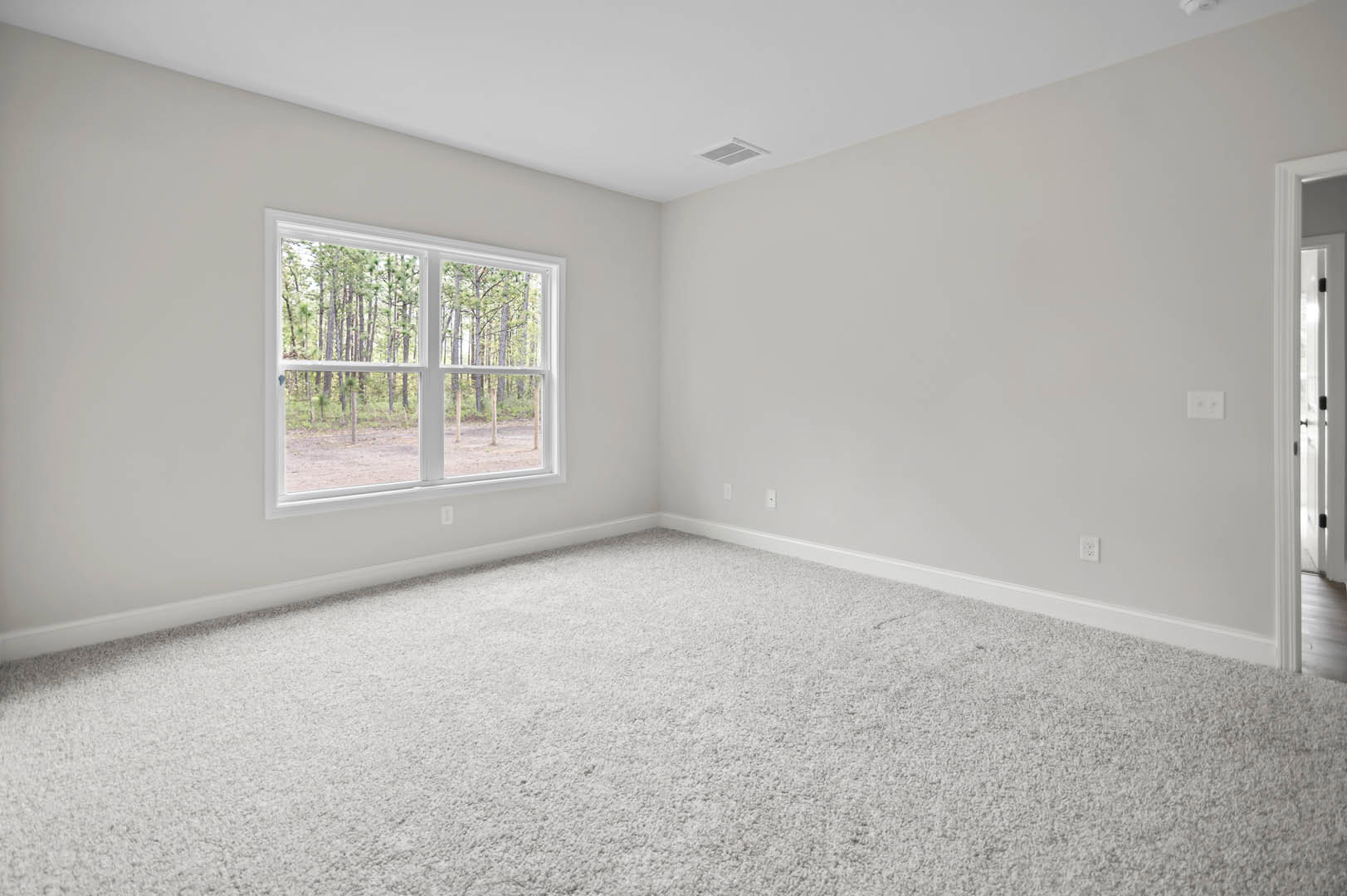 Bright room with white carpet, large window overlooking green trees, white walls, ceiling vent, and partially visible door