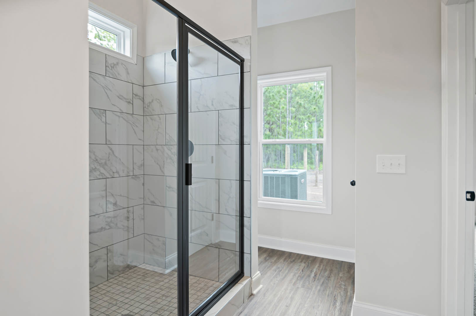 Bathroom featuring a glass shower enclosure, large window overlooking trees, white tile flooring, wall-mounted air conditioner, and modern light switches.