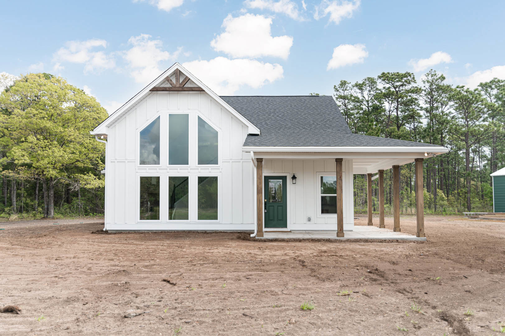 White house exterior with multiple windows, two green doors with glass panes, surrounded by dirt field and leafy tree under cloudy sky