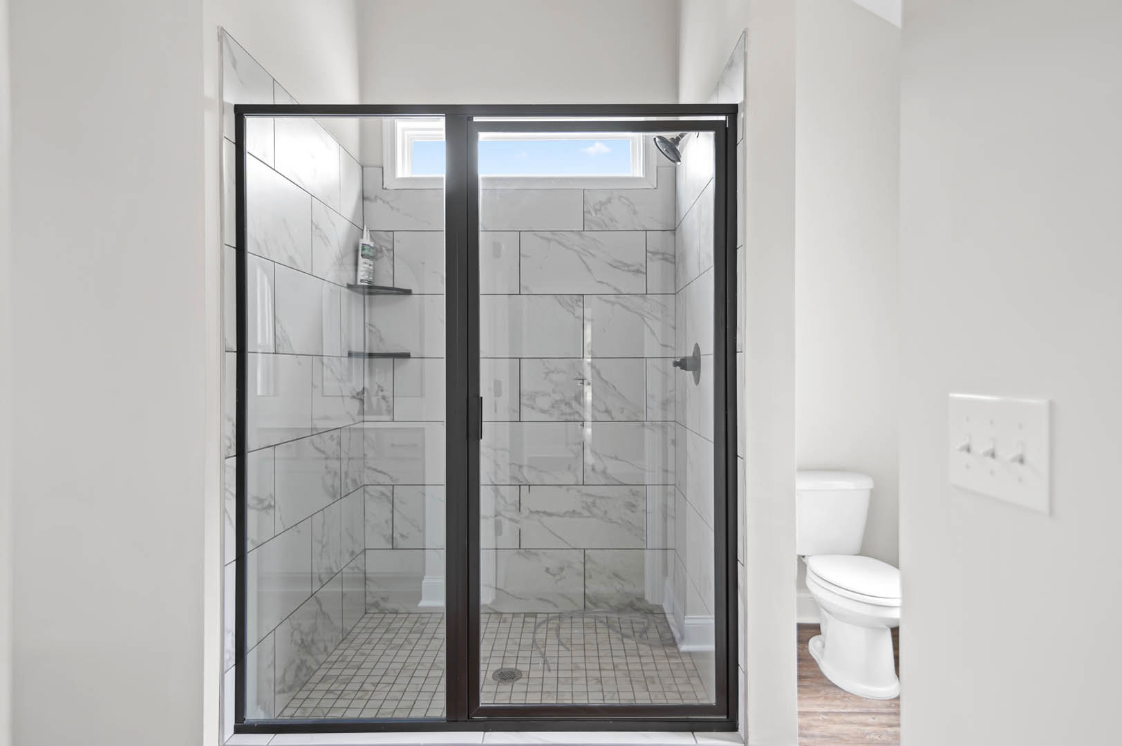 Walk-in shower with clear glass doors, large wall tiles, chrome fixtures, and partial view of adjacent toilet and light switch