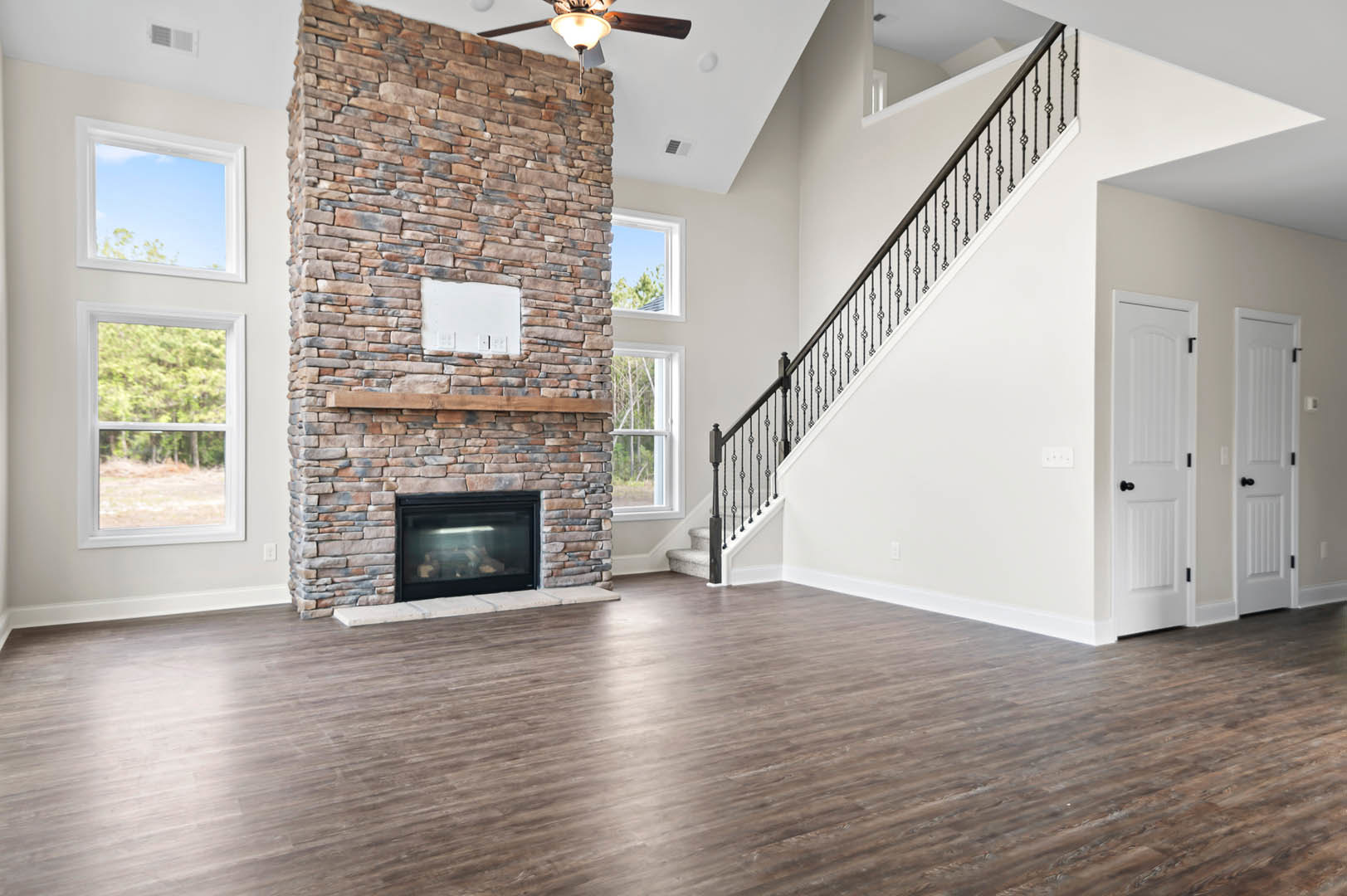 Living room featuring a stone fireplace with a glass-front insert, hardwood flooring, a staircase with black railing, large window overlooking trees, and a stone accent wall with