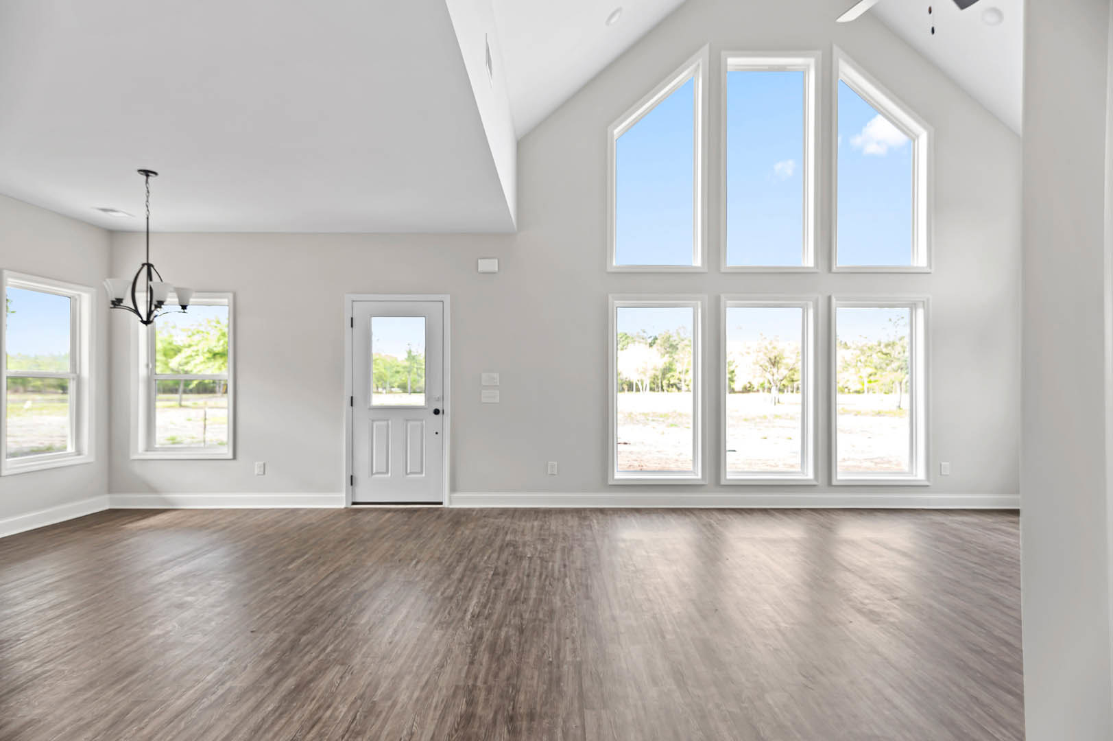 Spacious interior with light wood flooring, white trim, multiple large windows showing green trees outside, and a white door with glass panel