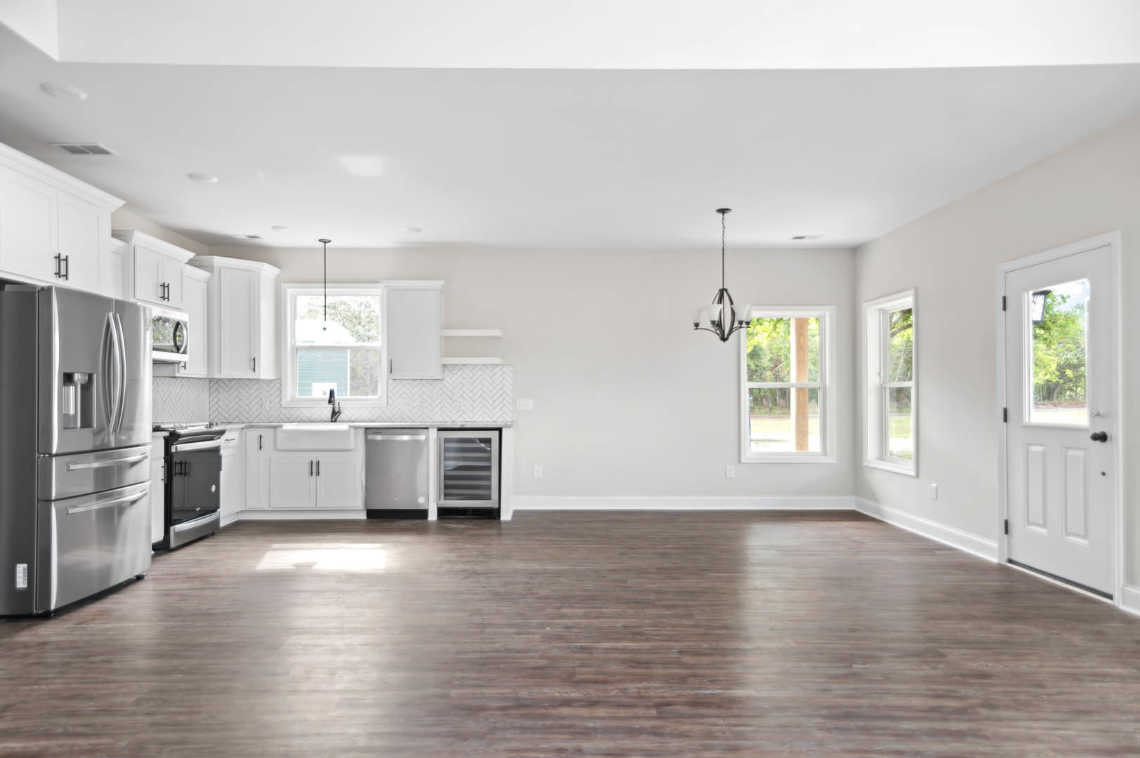 Open-concept kitchen and living room featuring hardwood floors, stainless steel refrigerator, white cabinetry, black and white pendant lamp, large window with white frame, and