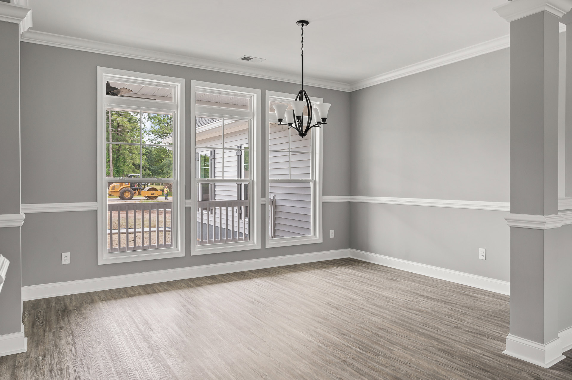 Wood flooring and white walls in a bright room with large windows, five-shade chandelier hanging from ceiling, view of trees and parked car outside