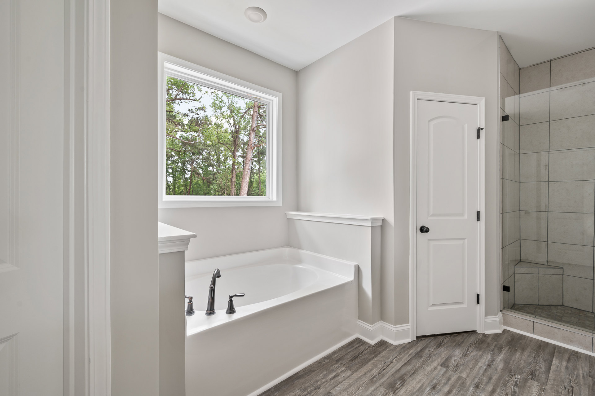 Freestanding white bathtub with chrome faucet beside large window overlooking trees, glass shower enclosure, white door with black hardware, light tile flooring