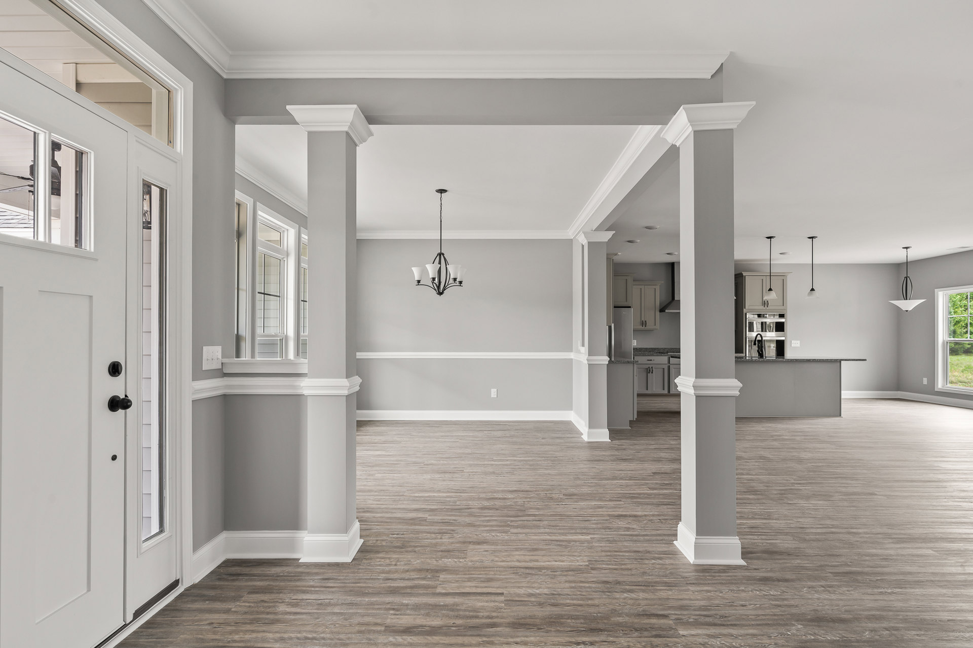 Open room with white columns, wood flooring, white trim, chandelier with white shades, large window showing outdoor view, and white door with black knobs