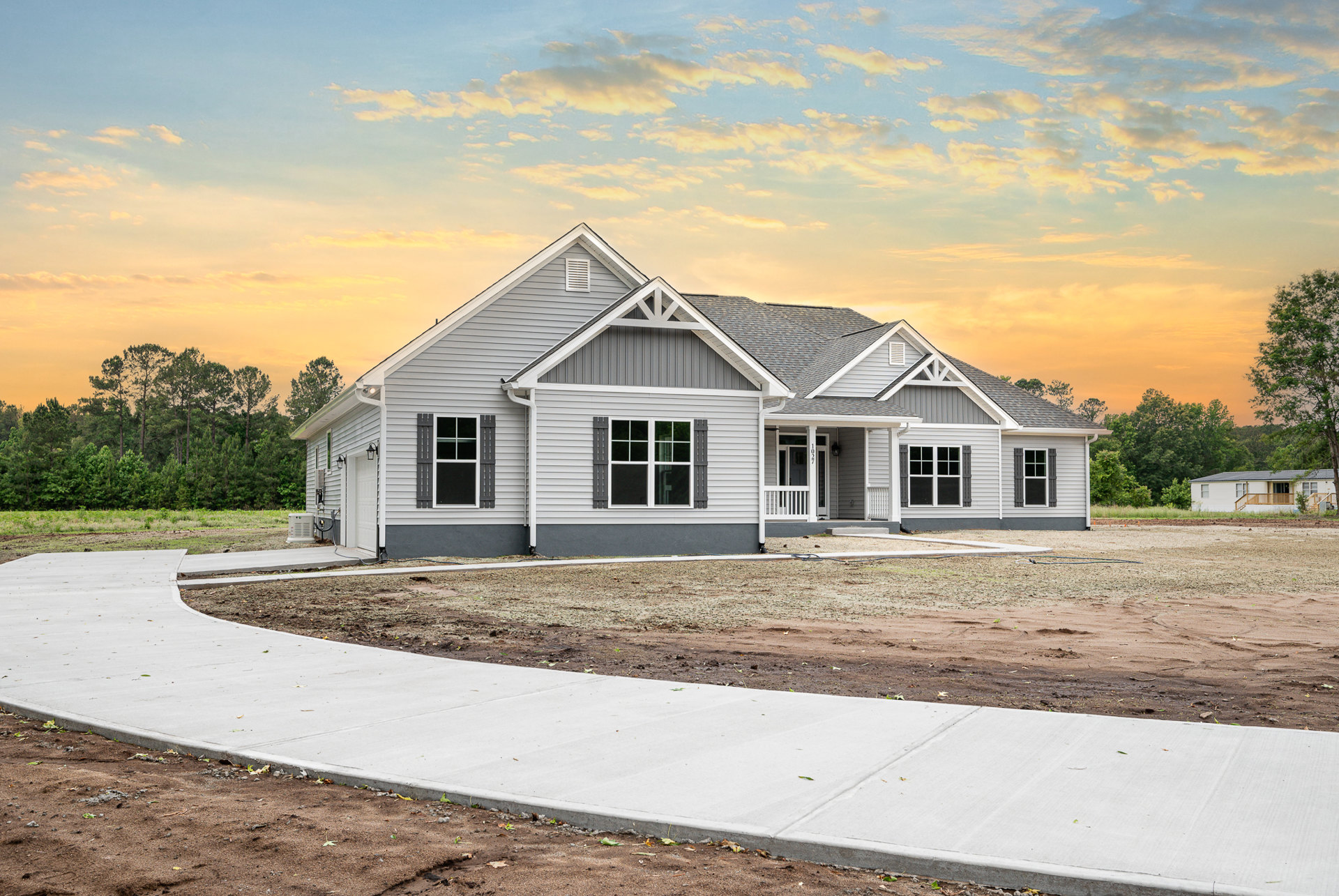 Two-story home with gray siding, white trim windows, covered front porch, concrete driveway, and landscaped yard with young trees under a cloudy sky