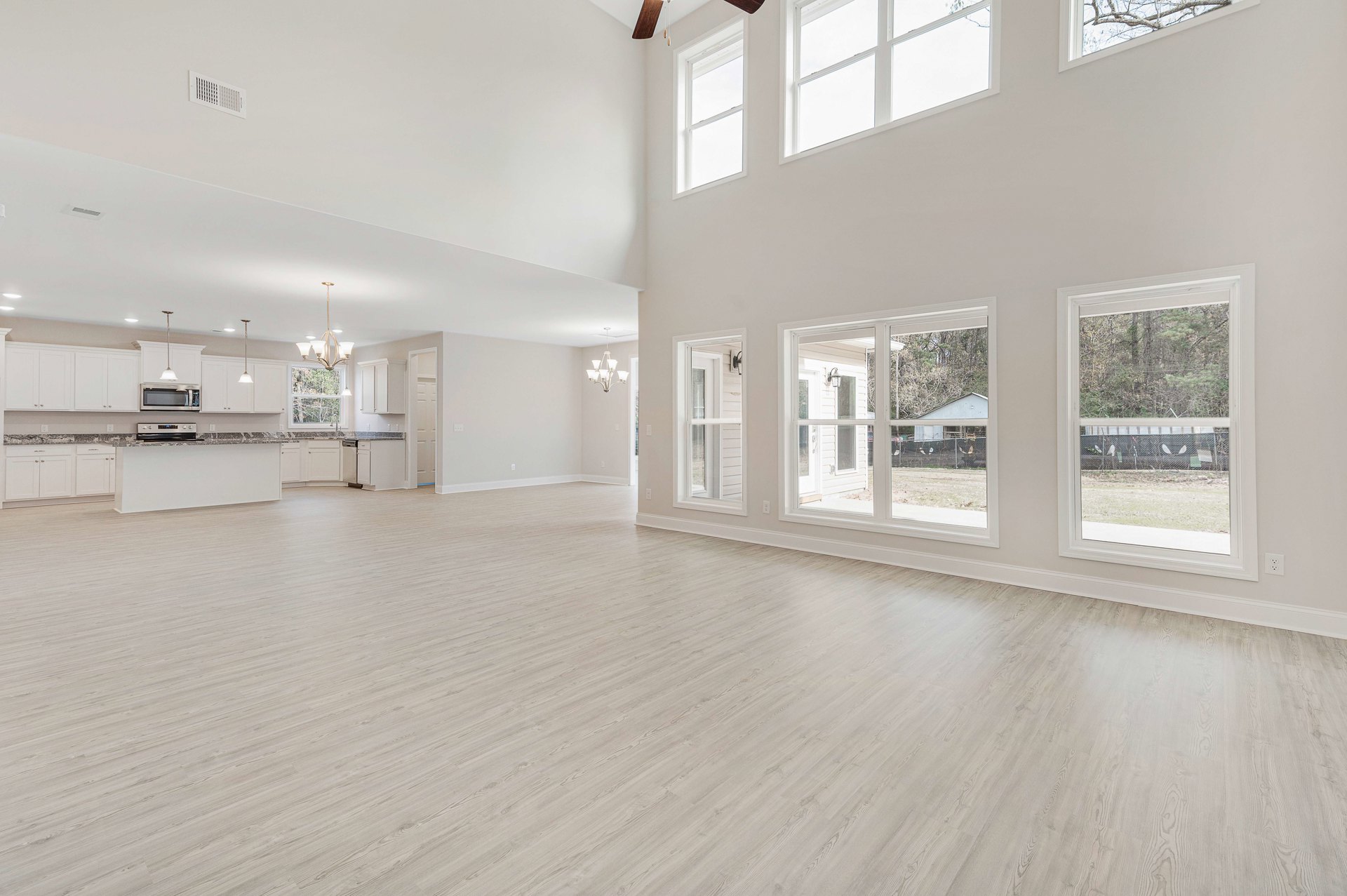Spacious room with white plaster walls, wide windows framed in white, light wood laminate flooring, and a chandelier with white shades hanging from the ceiling