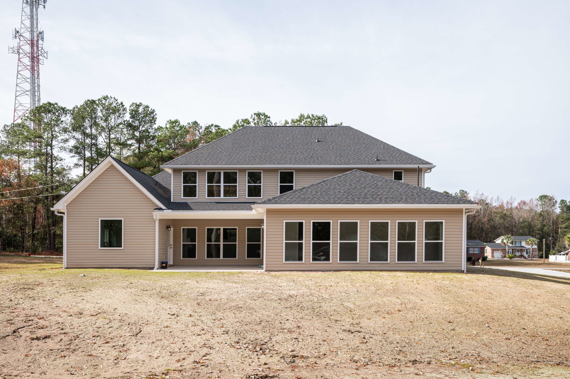 Two-story home with white-framed windows, illuminated interior, expansive dirt yard scattered with leaves, covered porch, mature trees, and cloudy sky overhead