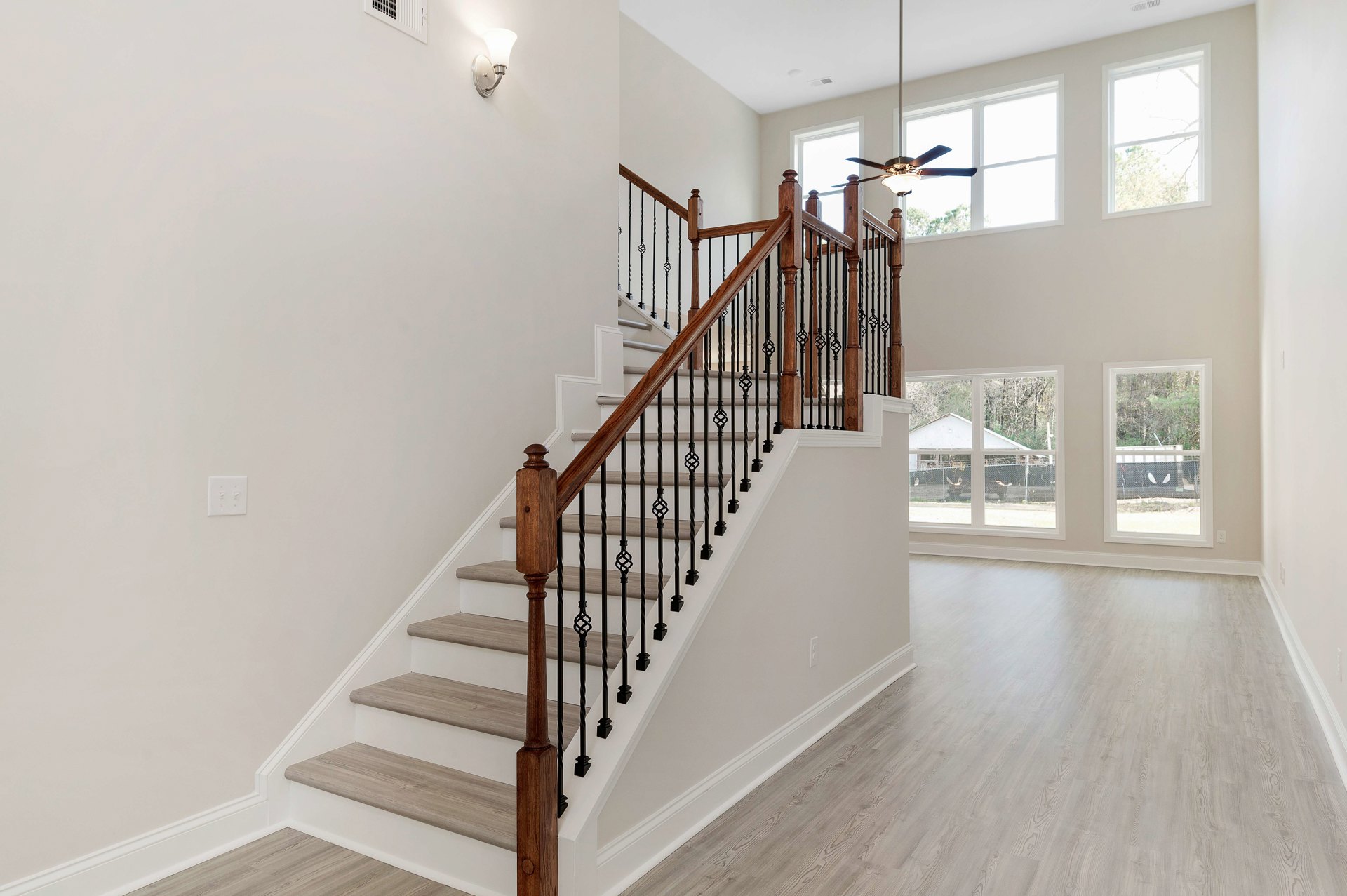 Wood staircase with matching railings and balusters, white-framed window overlooking fenced yard with trees, plaster walls, wall lamp, and light switch visible in foreground