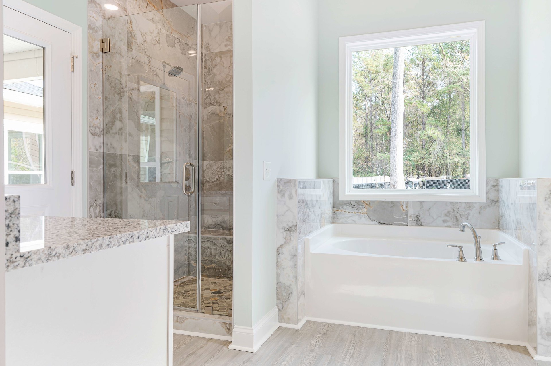 Bathroom with glass-enclosed shower, freestanding white bathtub with silver faucets, marbled countertop, large window overlooking trees, light tile walls and floor.