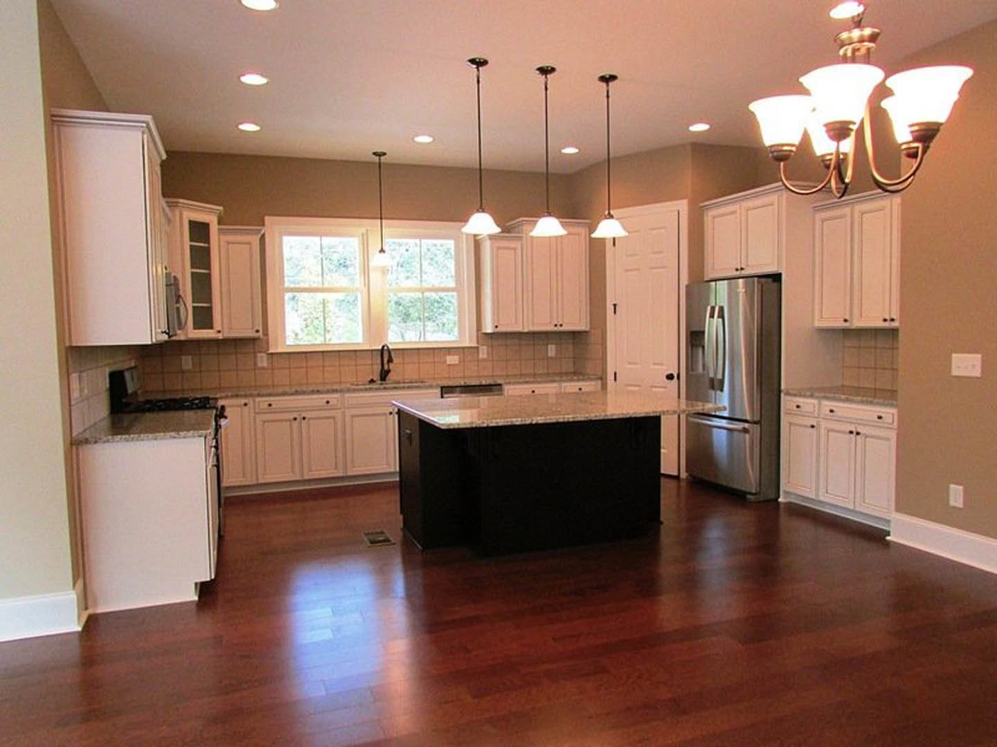 White cabinetry and black island with black countertop, stainless steel refrigerator with glass door, hardwood flooring, window, and modern chandelier in a contemporary kitchen.