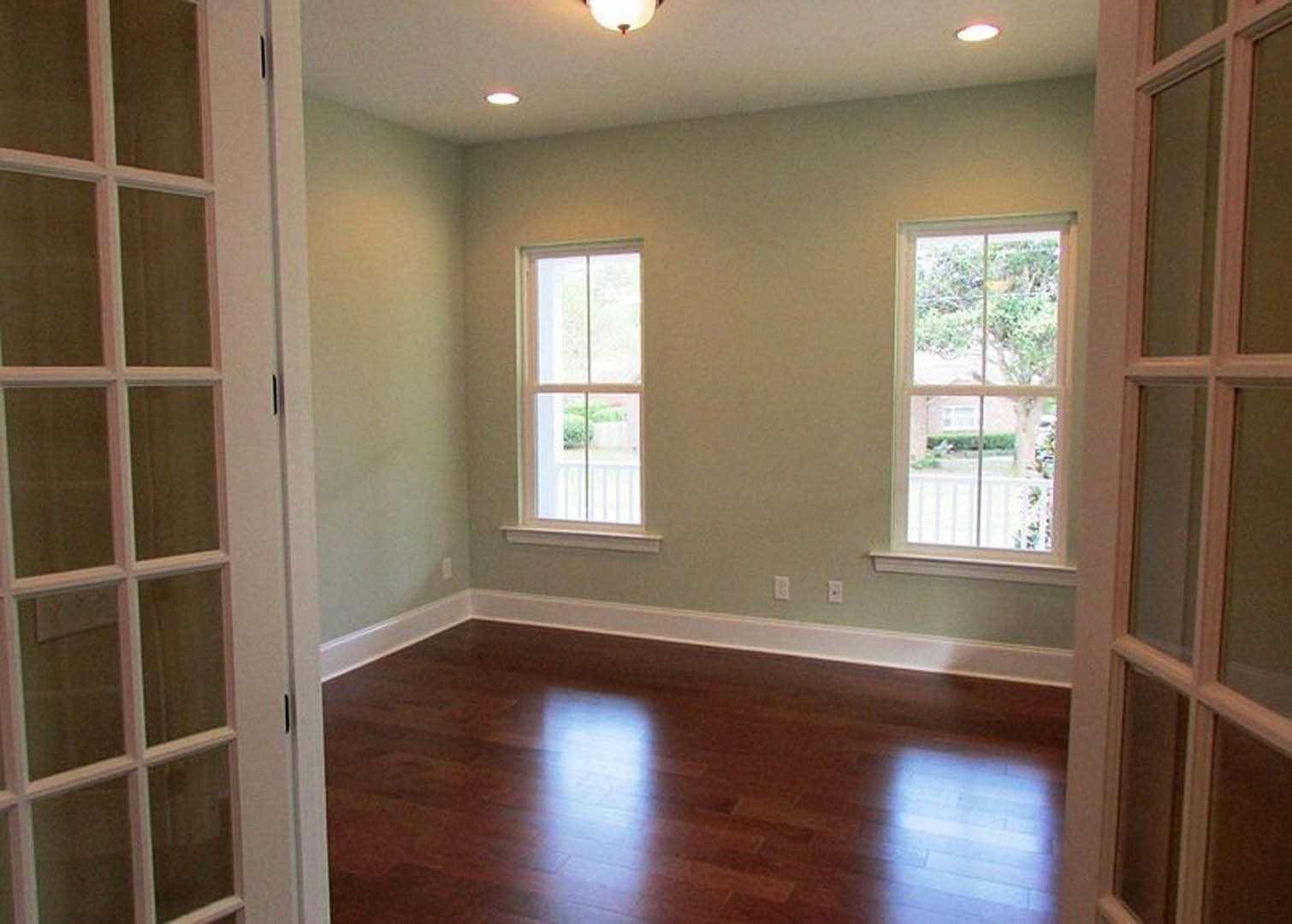 Sunlit room with polished hardwood floor, large windows overlooking green trees and bushes, and a ceiling light fixture