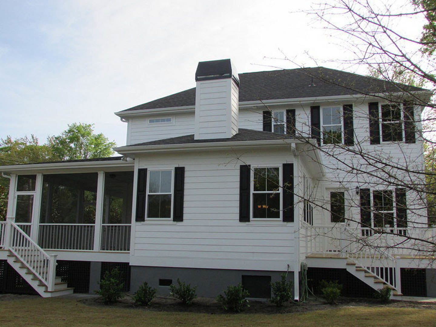 White siding house with black shutters, covered front porch, white staircase and railings, large windows with white frames, green landscaping and trees in background.