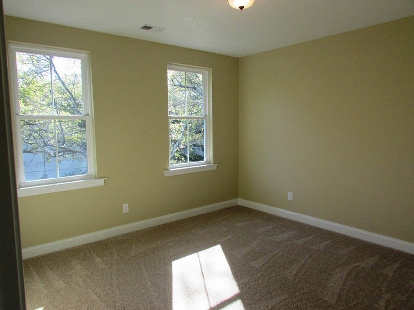 Beige-walled room with large windows, carpeted floor illuminated by natural light, view of green trees outside
