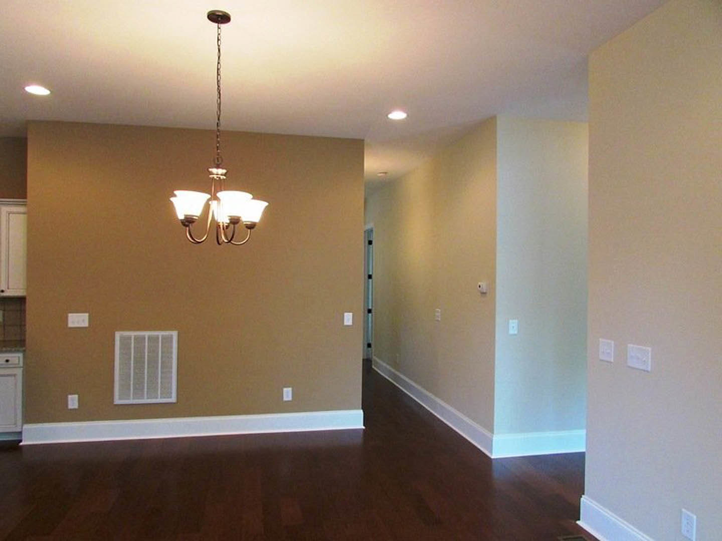 Hallway with dark wood flooring, white baseboards, plaster walls, and a ceiling-mounted chandelier; visible white electrical outlet and vent.