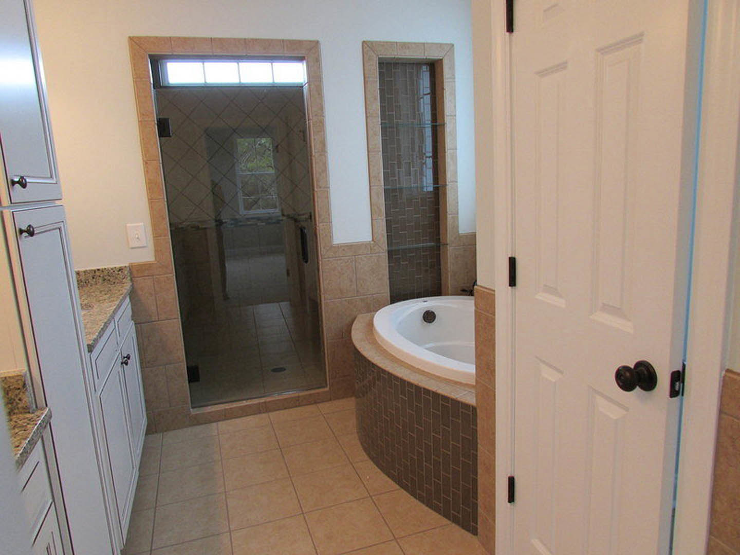 White bathtub with chrome faucet next to glass shower enclosure, light gray tile floor, white walls, and a brushed metal door knob.